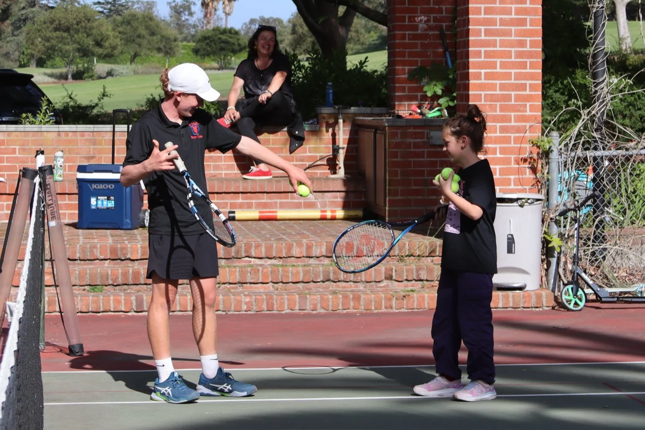A coach and student holding tennis rackets and tennis balls, playing tennis on an outdoor court with a woman sitting on the steps watching behind them.