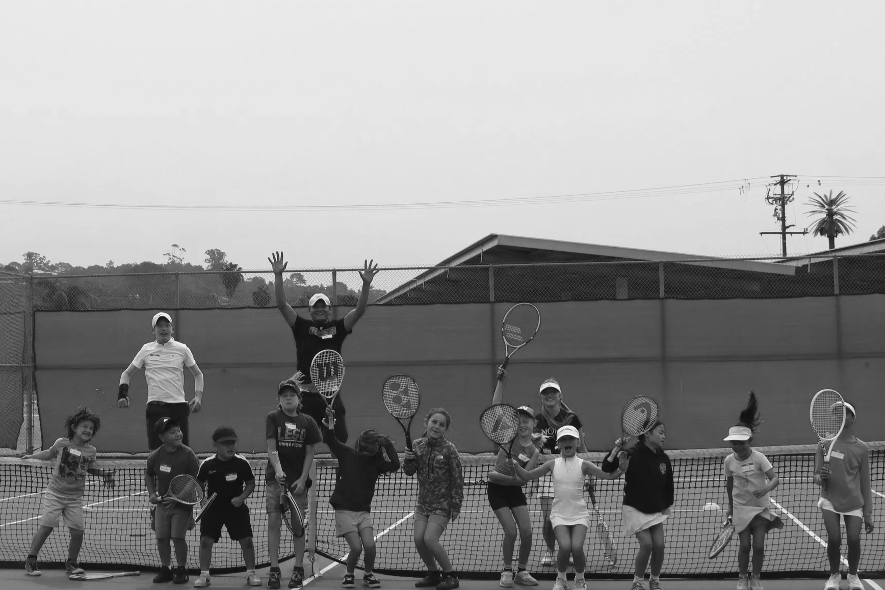 A group of children and a coach on a tennis court during a tennis practice or event. The coach is standing in the center, smiling with arms raised, holding a tennis racket. The children are holding tennis rackets, some appear excited and cheerful, with tennis balls and equipment visible. The background shows a fence, a building, and trees.