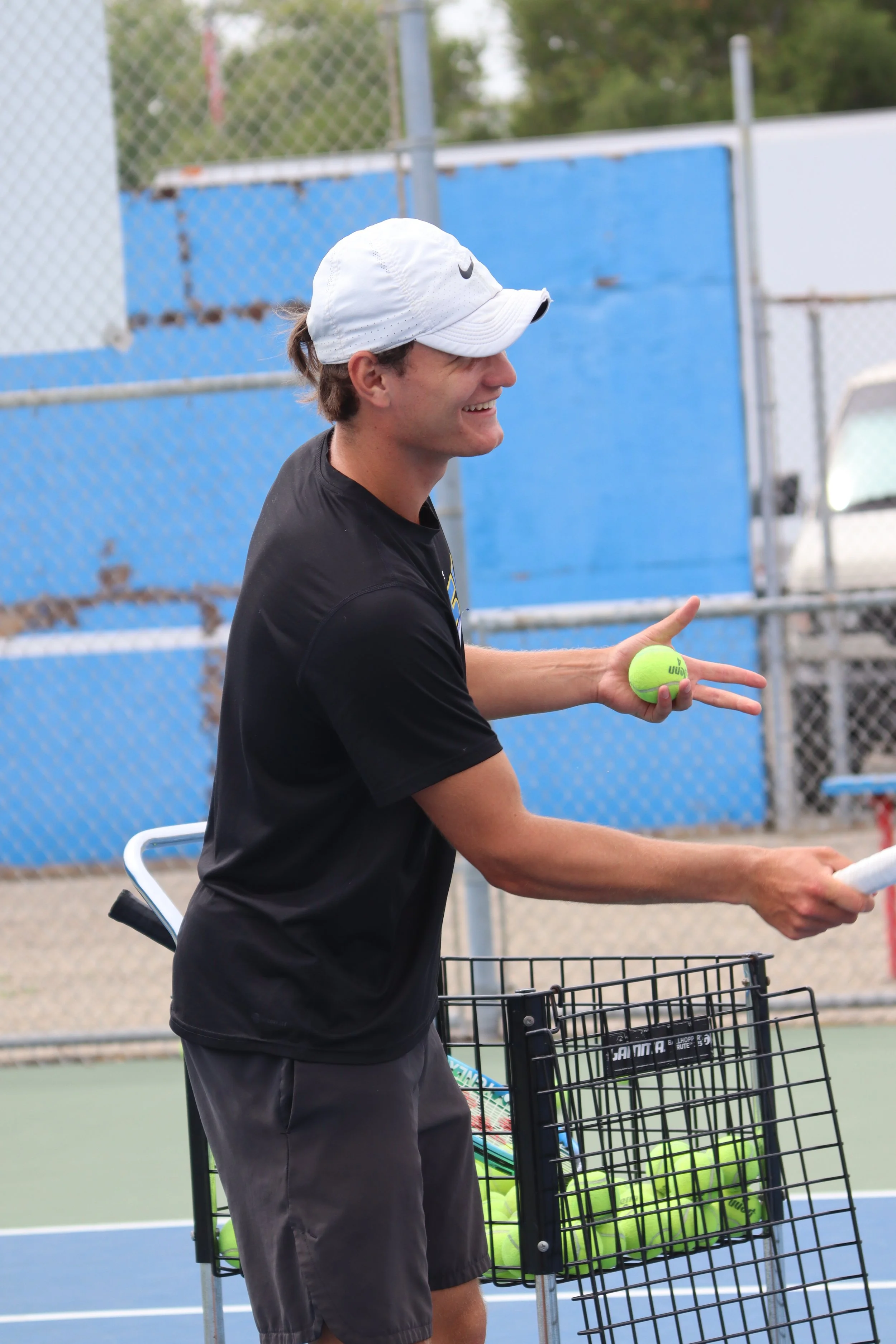 A coach in a black t-shirt and shorts holding a tennis ball while standing next to a basket filled with tennis balls on a tennis court, wearing a white cap.