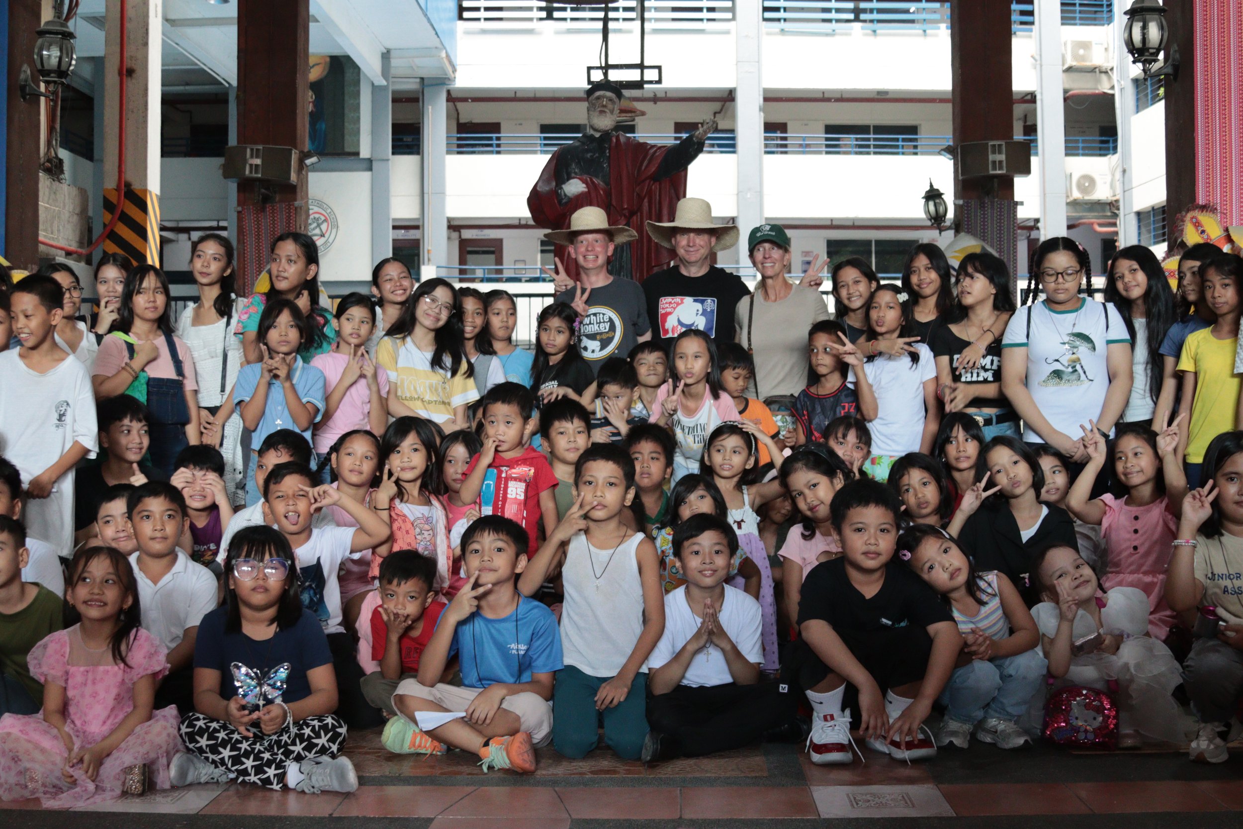A large group of children and a few adults posing for a group photo inside a building with a statue of a man in the background. The children are sitting and standing, some making peace signs or smiling at the camera.