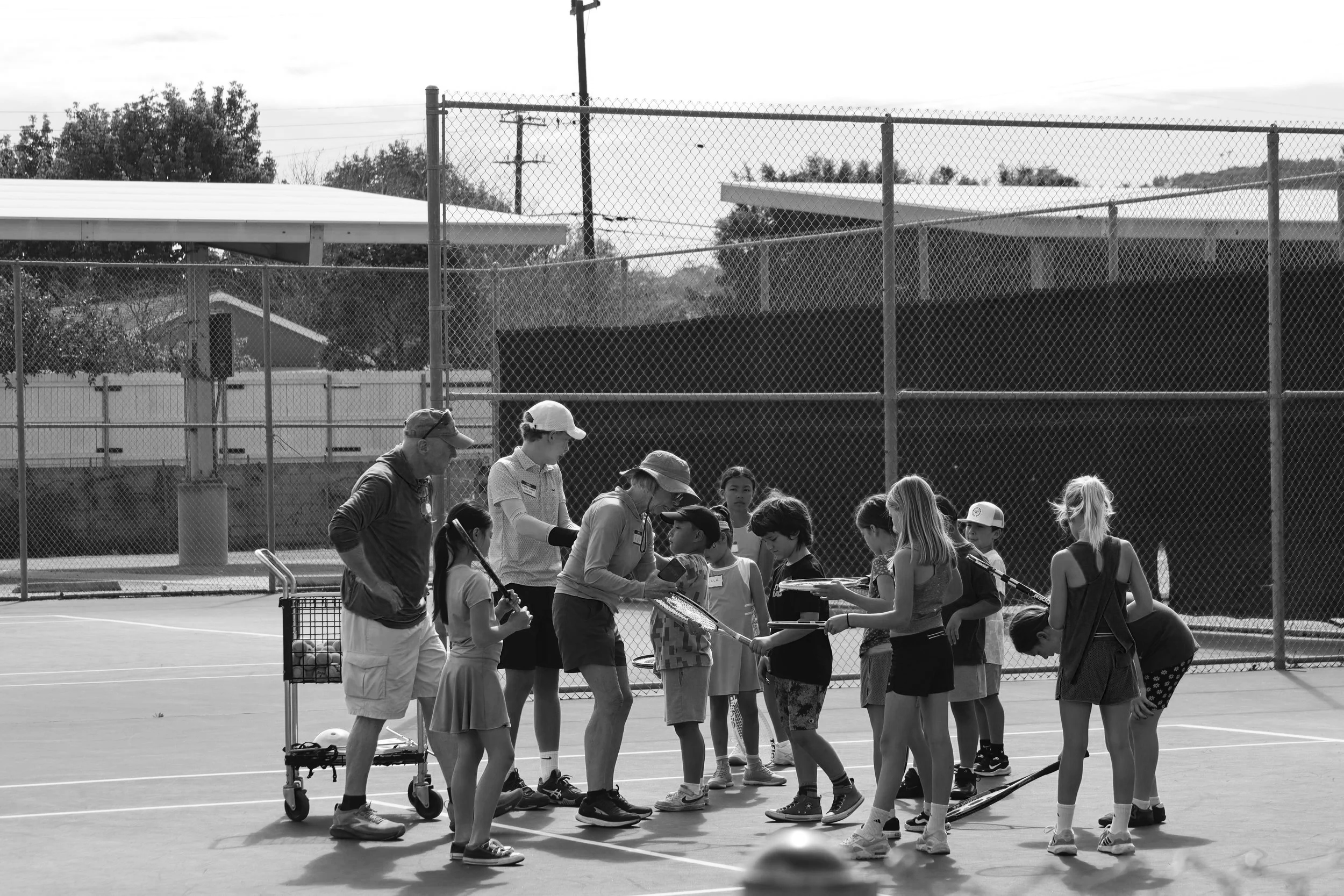 Children and adults standing in a line on a tennis court, chain-link fence, trees, and buildings in the background.