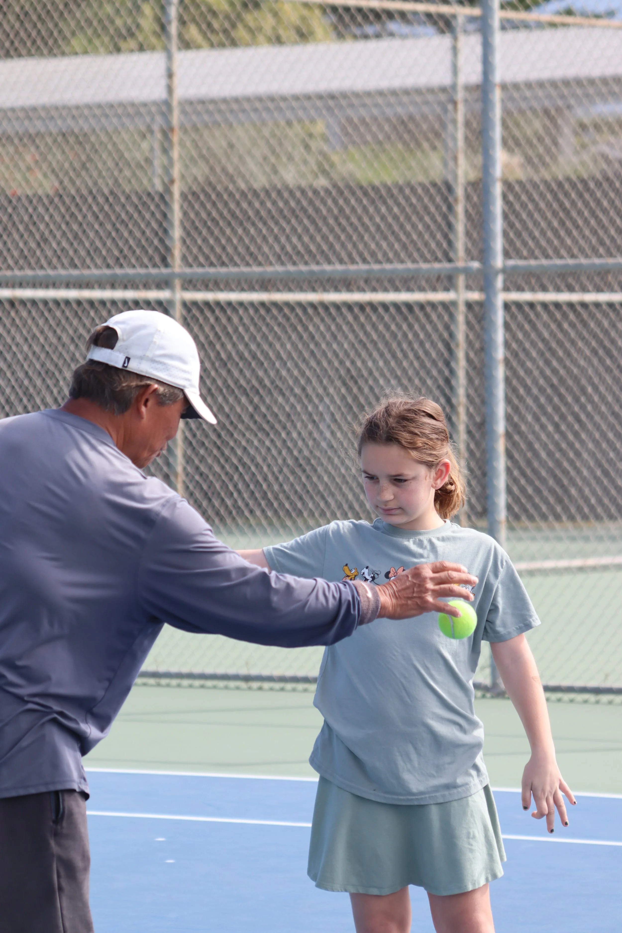 A girl and a coach on a tennis court, with the man feeding her a tennis ball during a coaching session. The court is outdoors with a chain-link fence in the background.