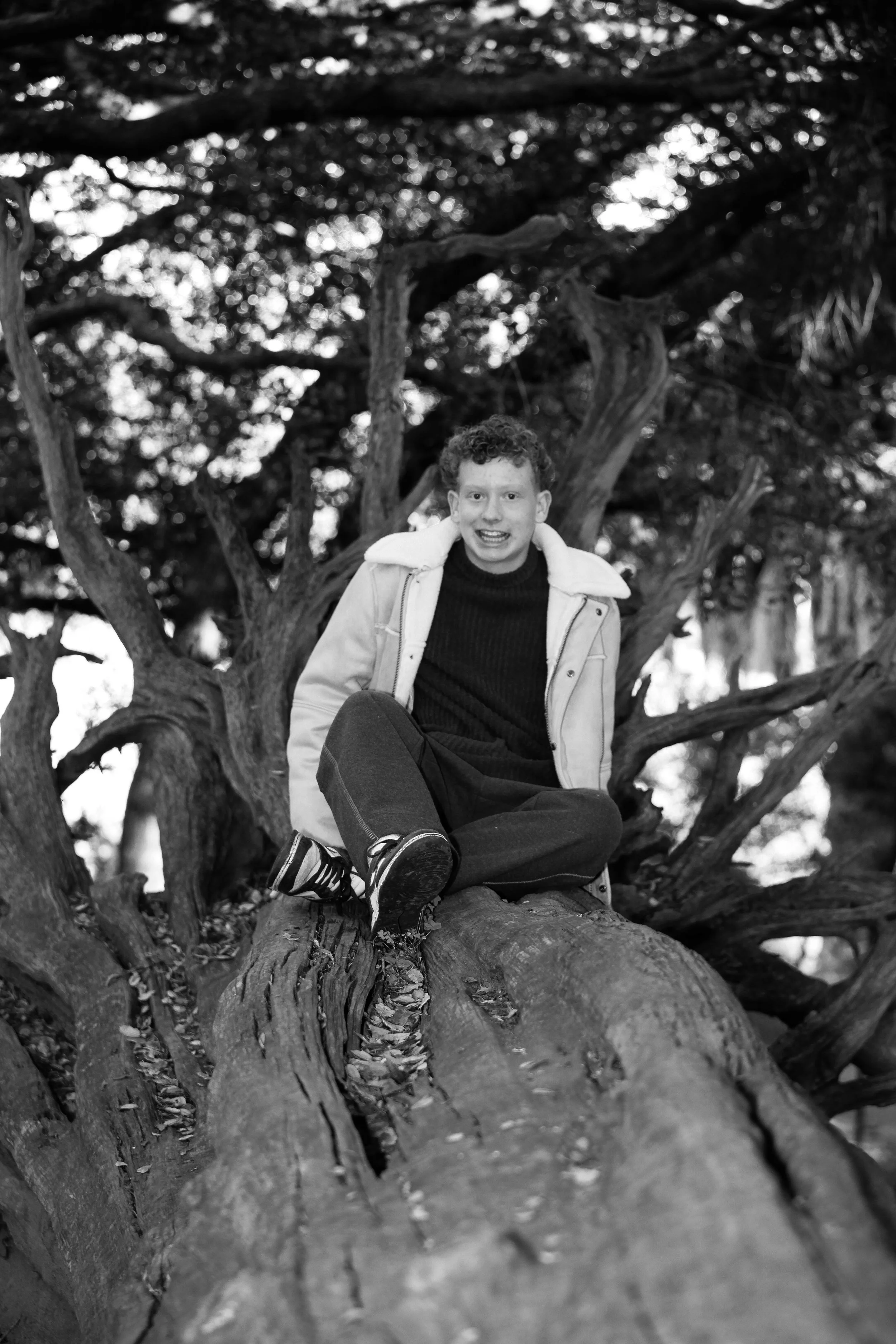 A young man sitting on a large tree branch, surrounded by the tree's twisting branches and leaves, smiling at the camera in a black and white photo.