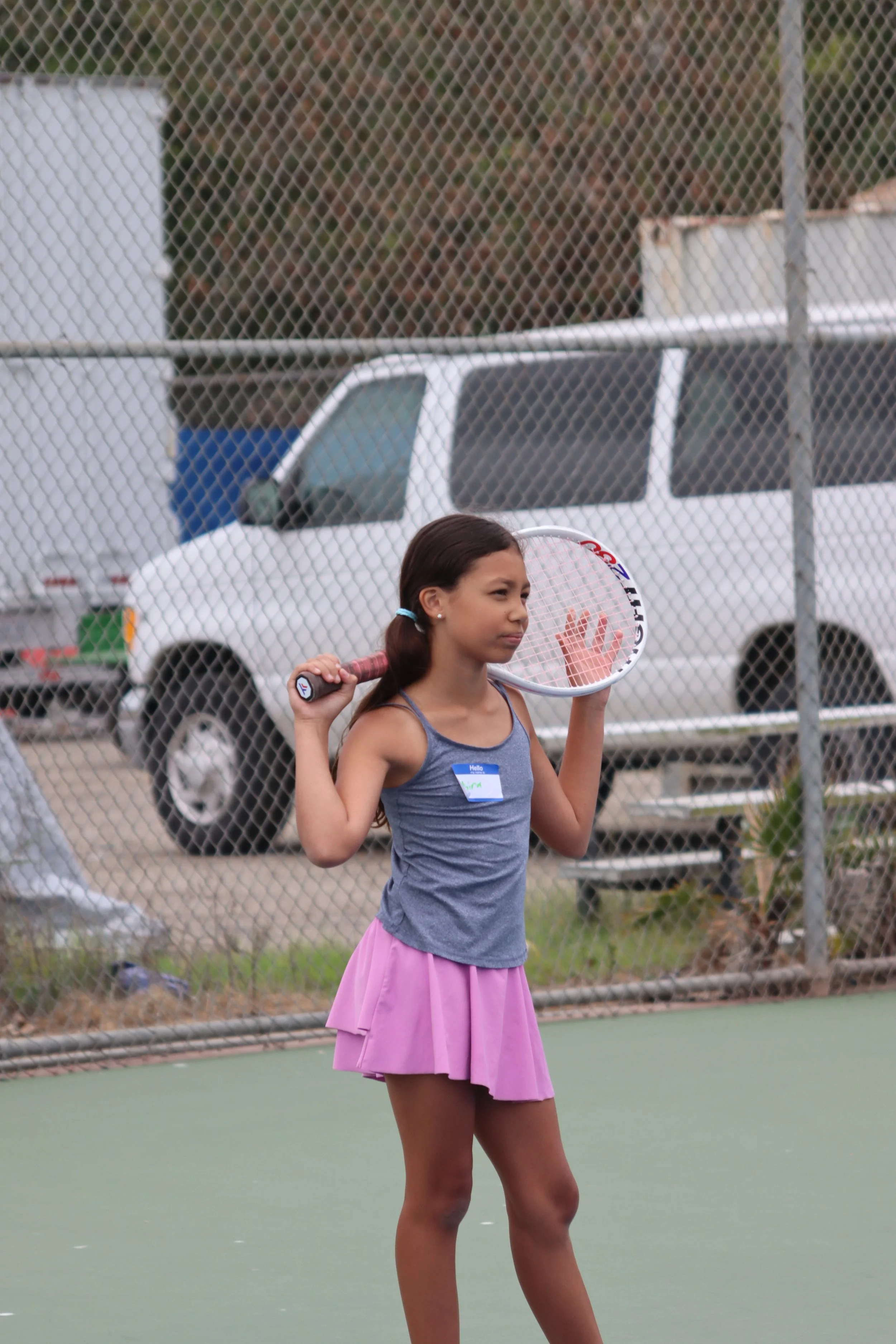 Young girl with a tennis racket standing on a tennis court behind a chain-link fence, with cars and trees in the background.