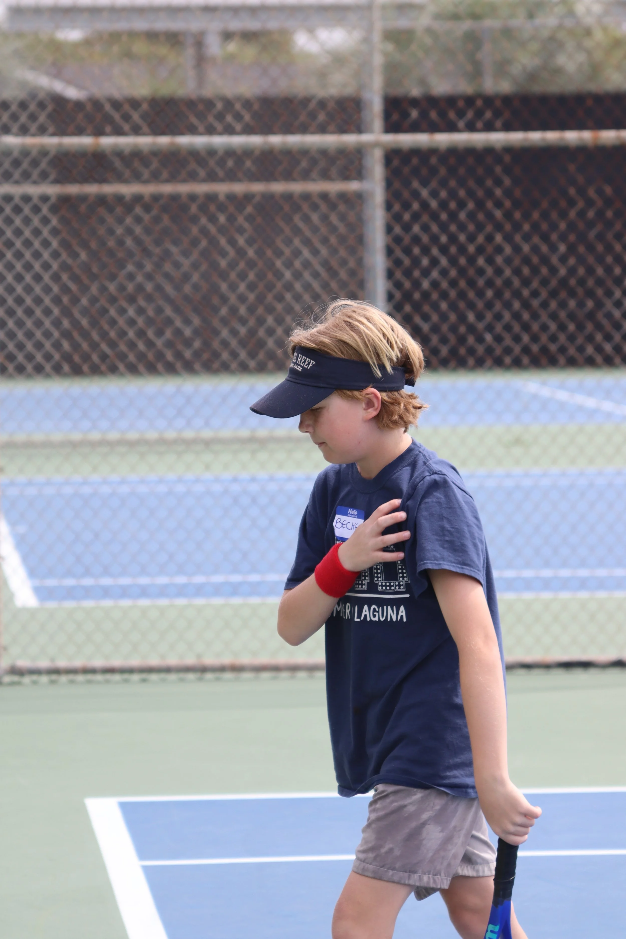 A young boy with blond hair wearing a navy blue golf visor, a navy t-shirt with the words 'MARI LAGUNA' and a name tag reading 'BECKET,' and khaki shorts, holding a tennis racquet while standing on a tennis court with a chain-link fence in the backgr