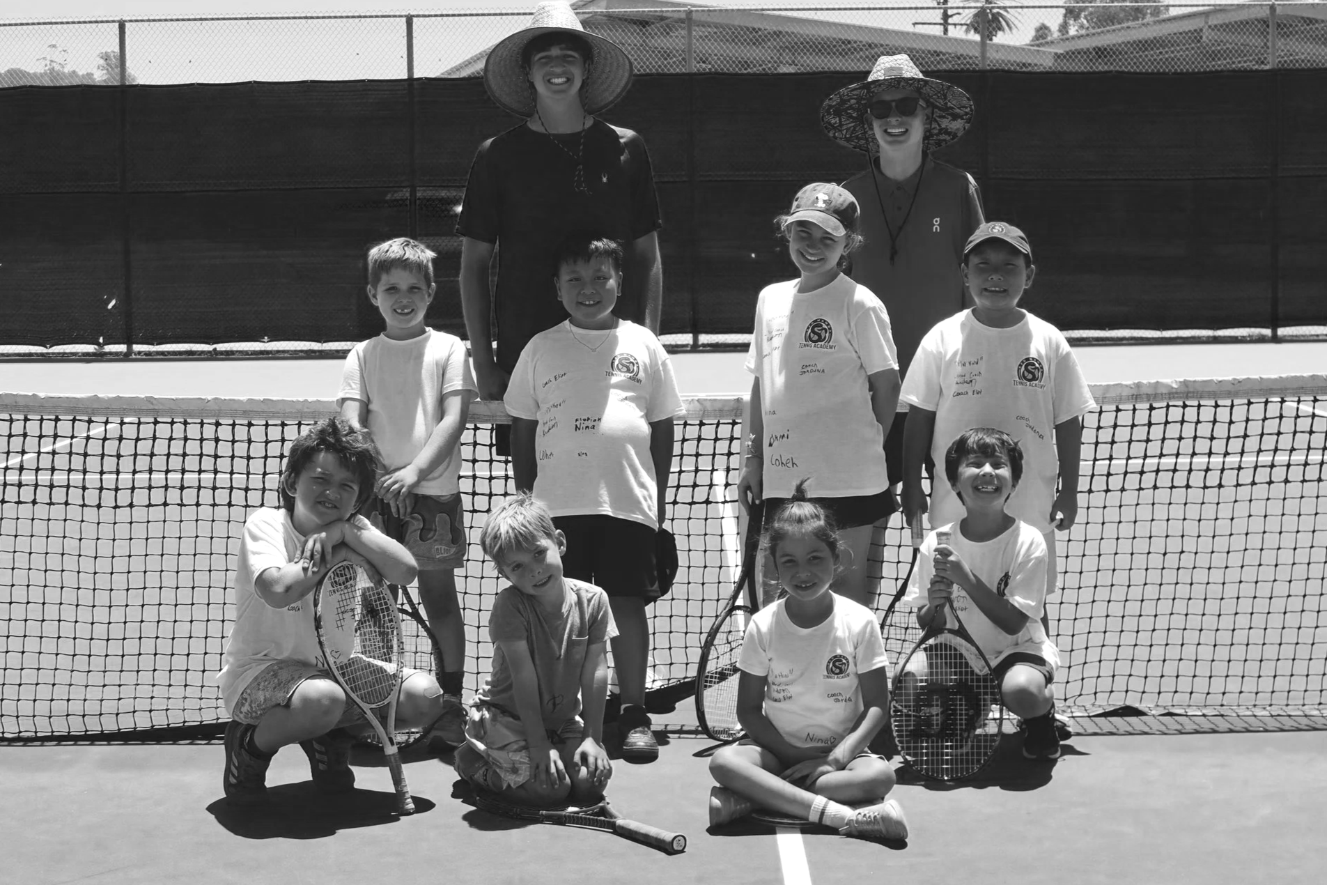 Group of children and two women on a tennis court, smiling, with tennis rackets, behind a tennis net and fence, sunny day.