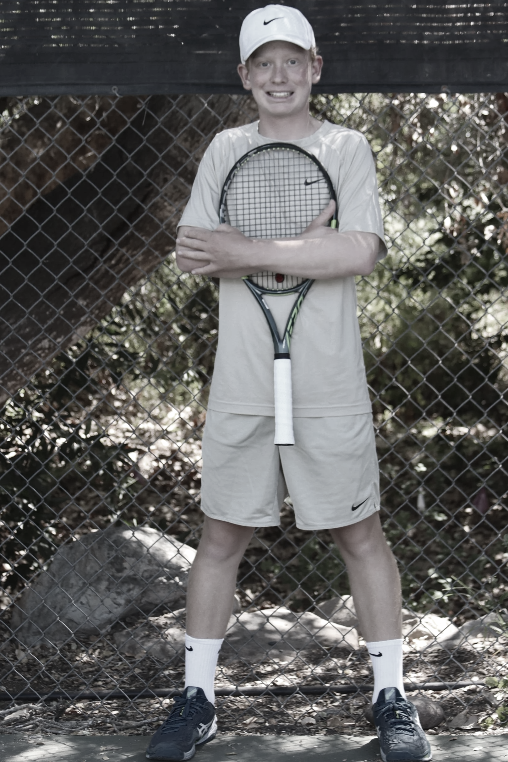 A young man dressed in athletic wear standing outdoors in front of a chain-link fence, holding a tennis racket.