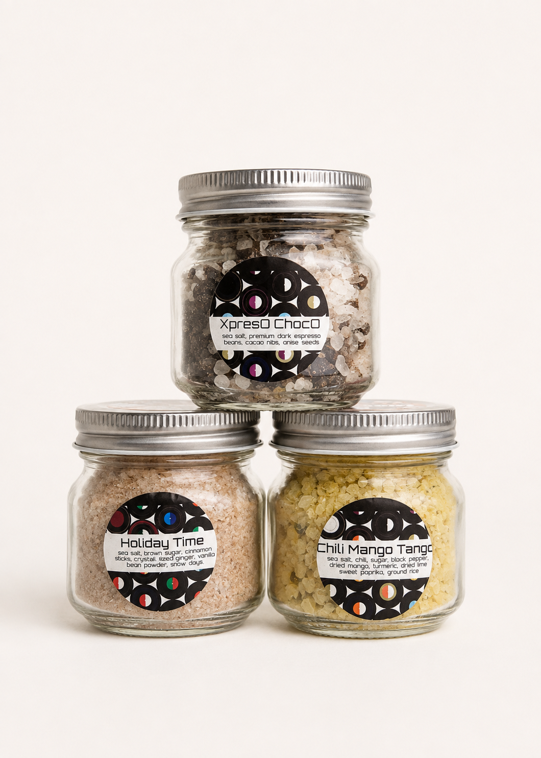 Three glass jars with metal lids containing colorful salts and seasonings, labeled Xpreso Choco, Holiday Time, and Chili Mango Tango, arranged in a pyramid on a white background.
