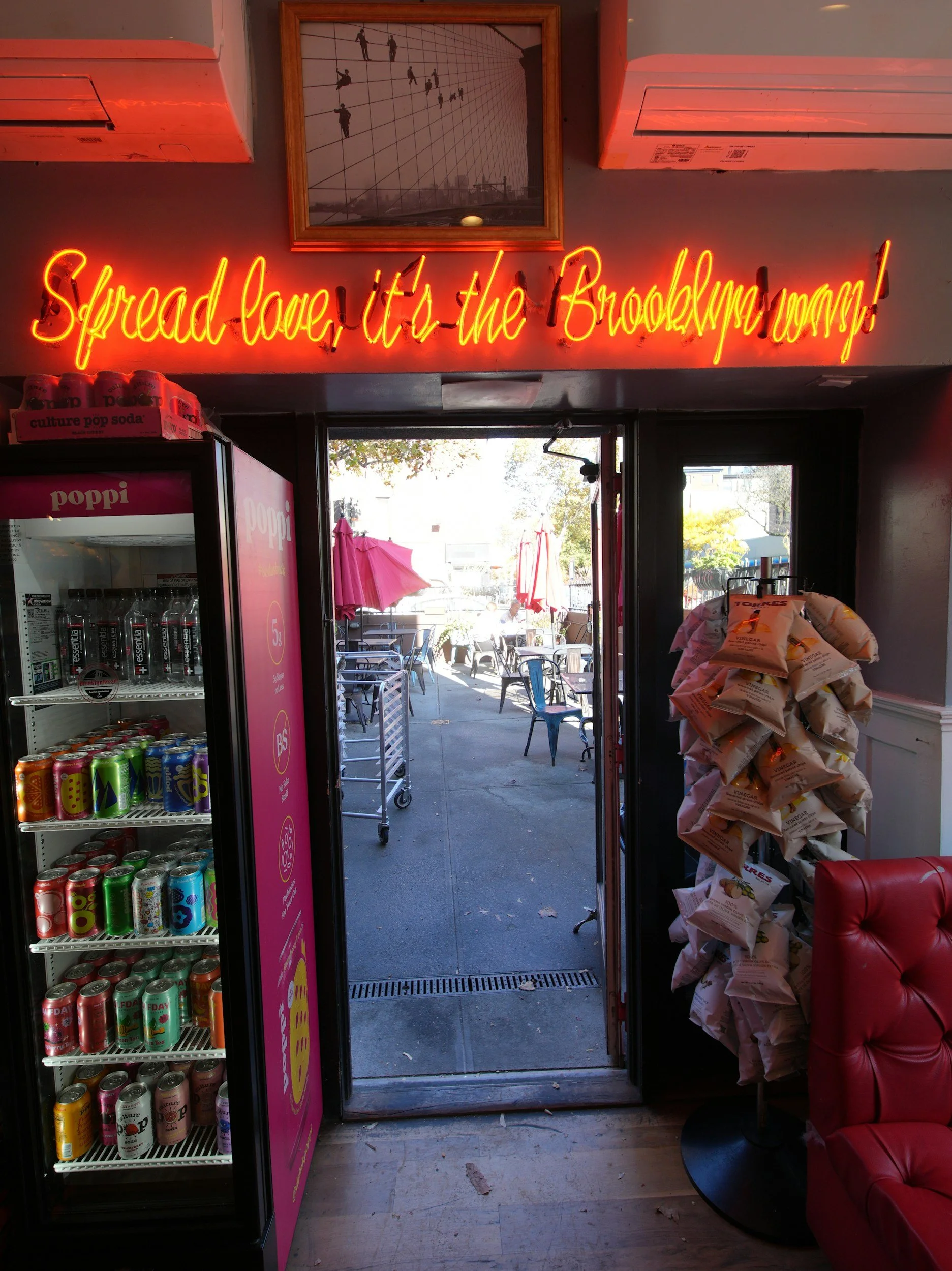 Entrance to a restaurant or cafe with a neon sign that says 'Spread love, it/'s the Brooklyn way!'. The door is open, showing outdoor seating with umbrellas. Inside, there is a vending machine with beverages, a pile of bags, and a red upholstered chair.