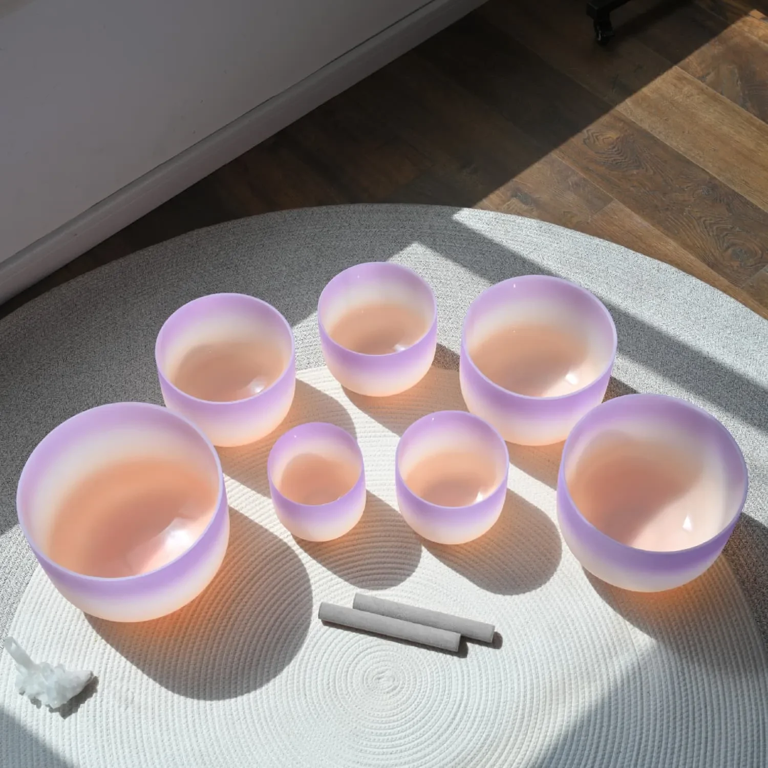 Seven pastel purple and pink singing bowls and two white sticks on a white textured mat with sunlight and shadow.