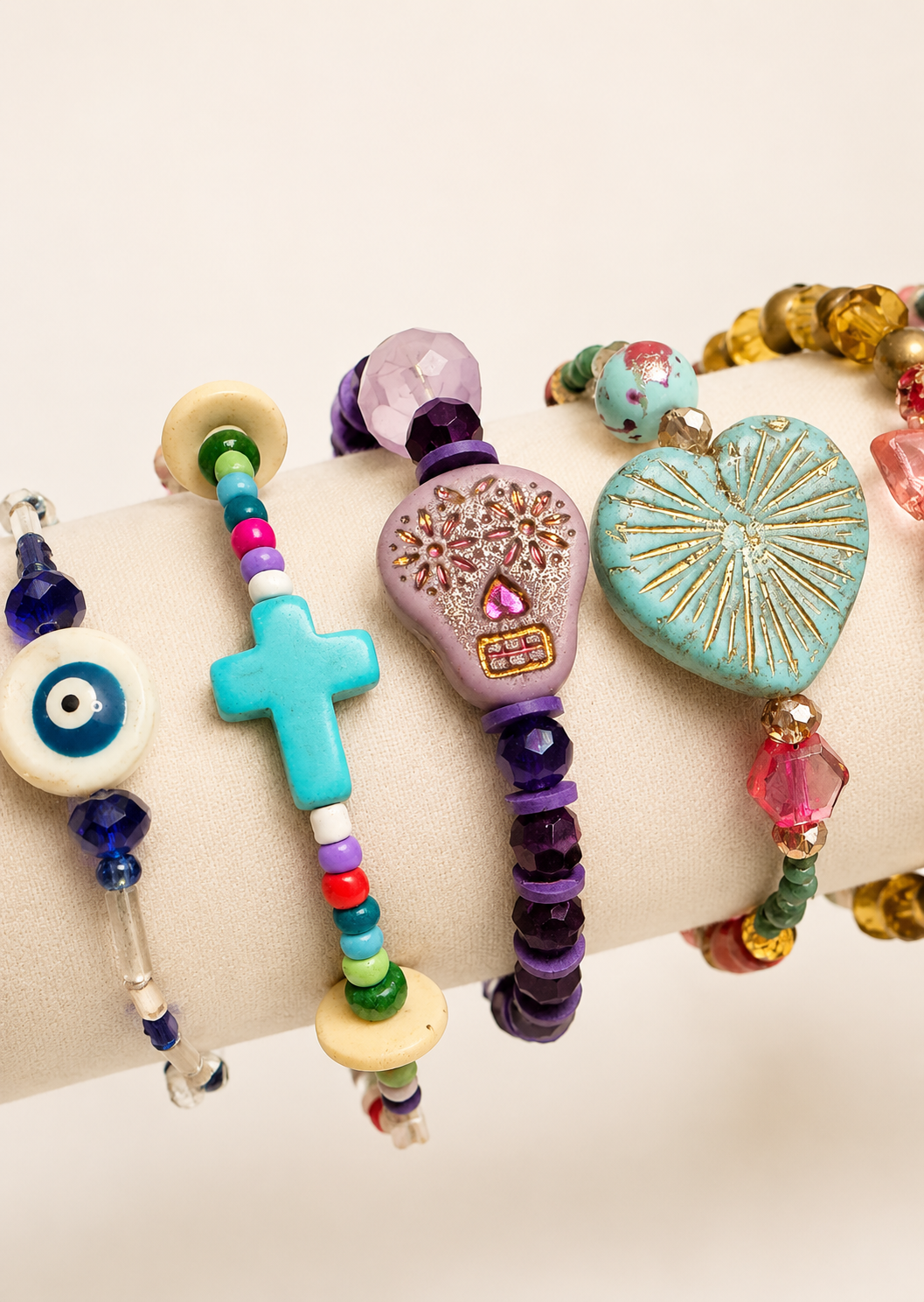 Colorful beaded bracelets displayed on a beige jewelry stand, featuring various charms and designs, including a blue cross, a purple skull, a turquoise heart, and an eye symbol.