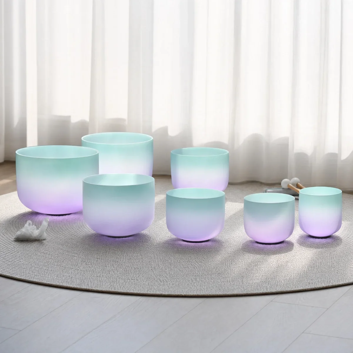 Set of seven pastel-colored crystal singing bowls on a circular rug in front of white curtains, with mallets nearby.