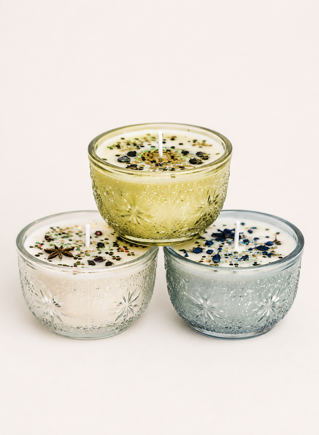Three decorative glass candles with white wax and multicolored sprinkles, arranged in a pyramid shape on a white background.