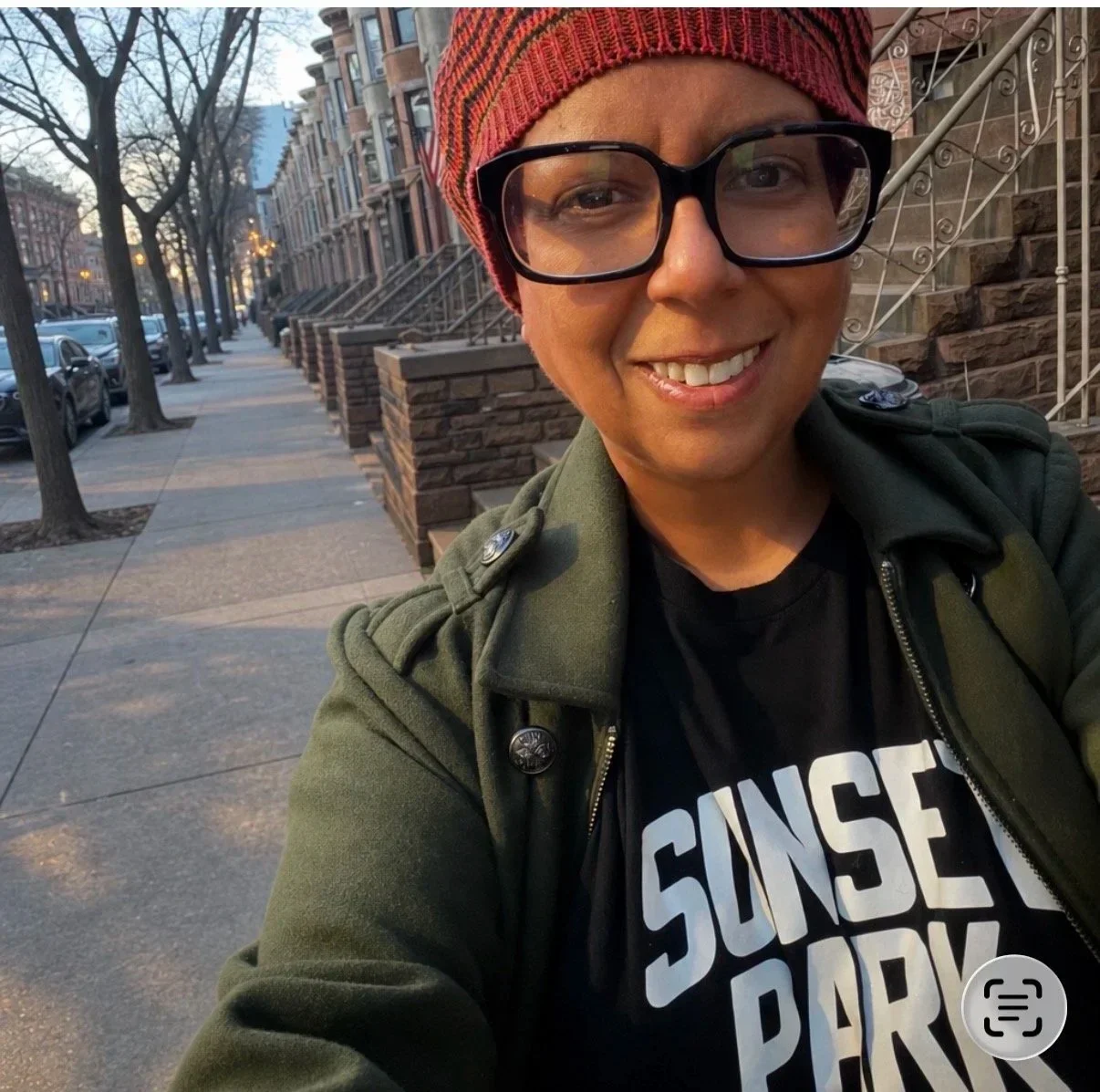 A woman taking a selfie on a city sidewalk during sunset. She is smiling, wearing glasses, a red knit hat, a green jacket, and a black T-shirt that says 'Sunset Park'. Brownstone buildings and parked cars line the street in the background.