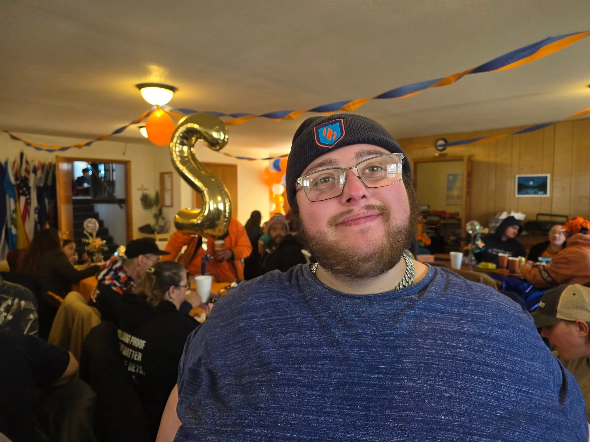 A man wearing glasses, a black beanie with an orange and blue logo, and a blue shirt, smiling at a party with orange and blue streamers, balloons, and people sitting and standing at tables inside a decorated room.