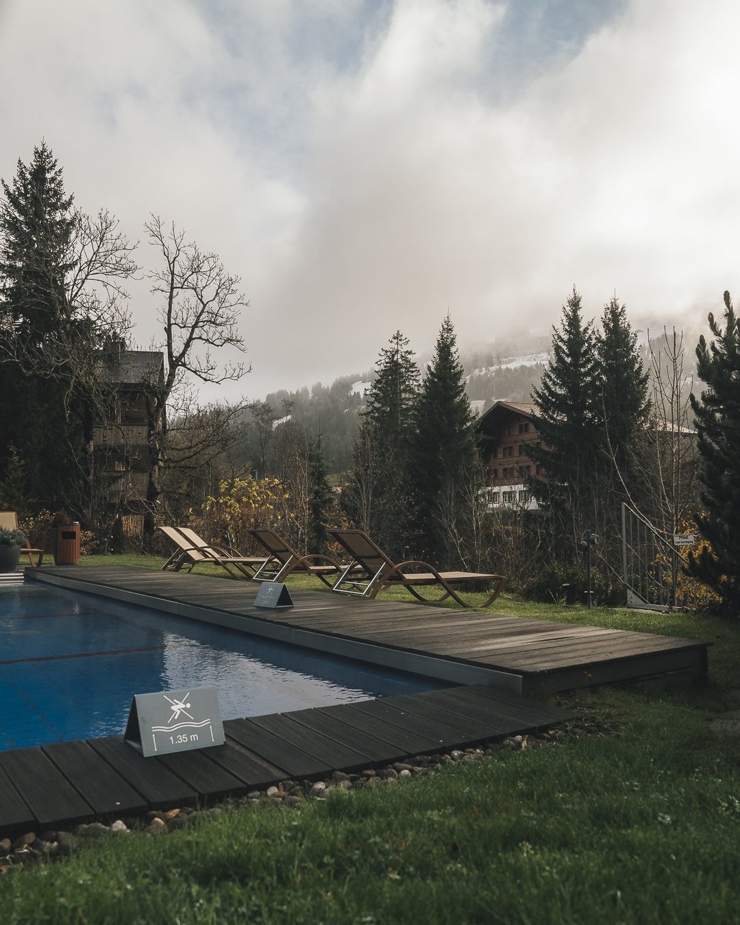 Ermitage Wellness & Spa Hotel in Gstaad, Switzerland. An outdoor swimming pool with a wooden deck and three lounge chairs, surrounded by trees, with mountains in the background and misty clouds overhead.