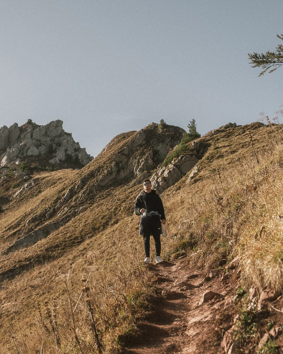 A person hiking on a narrow dirt trail on a hillside with dry grass, rocks, and sparse trees, with mountains in the background and clear sky above.