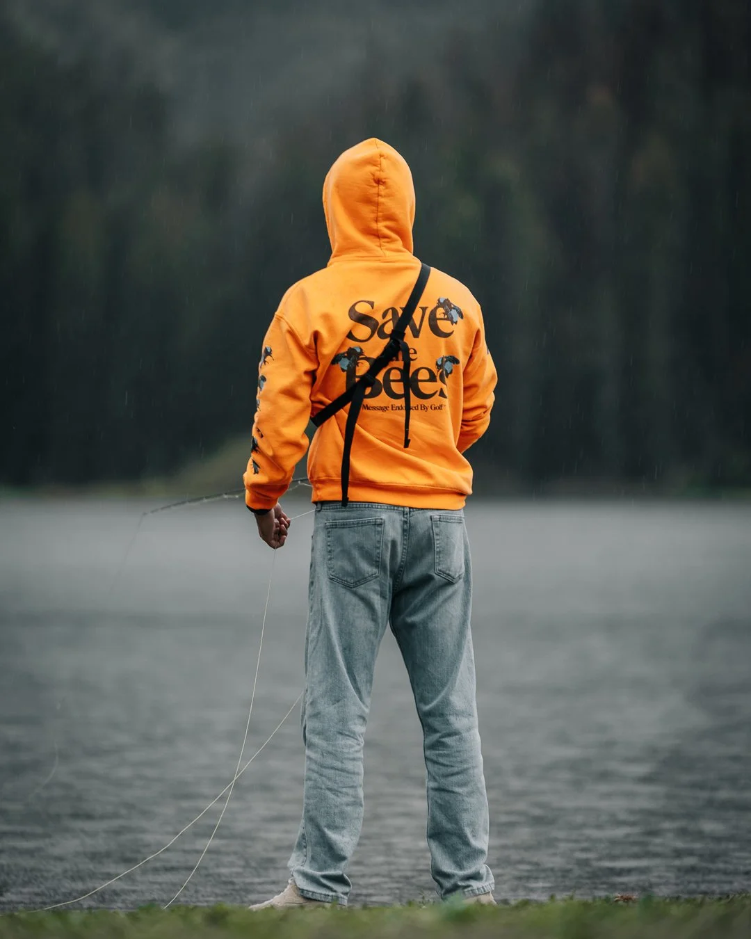 Person in an orange hoodie with 'Save the Bees' written on the back, standing by a body of water with their back to the camera, holding a fishing rod.