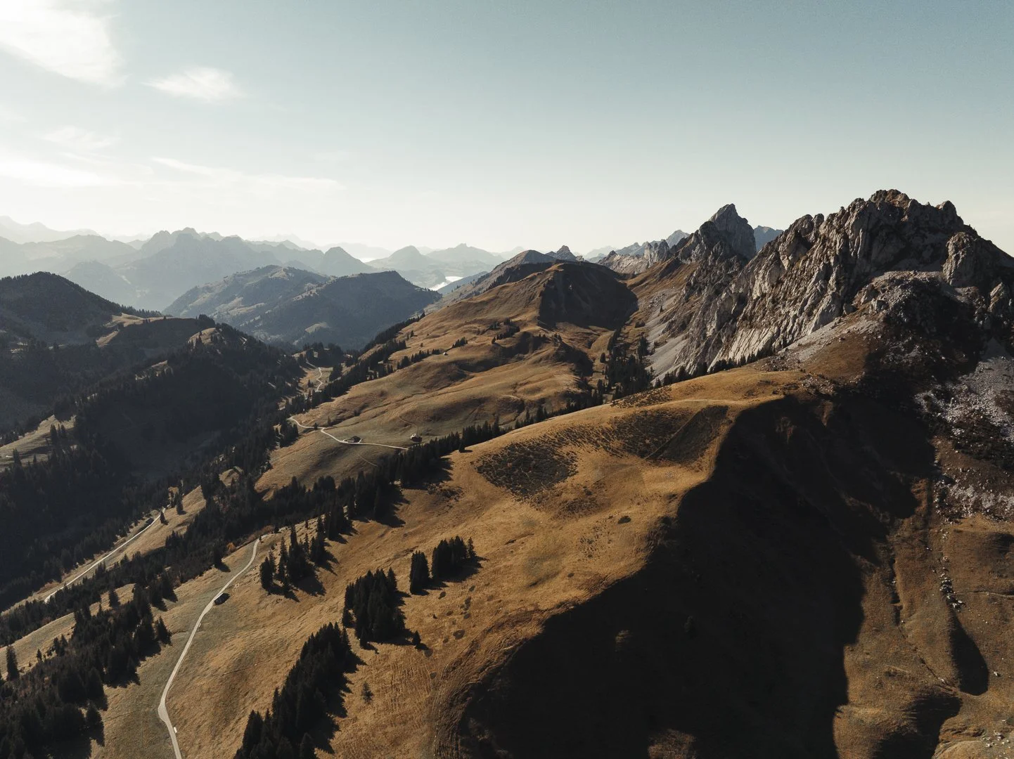Aerial view of rolling hills and rugged mountain peaks with a winding road through a mountainous landscape.