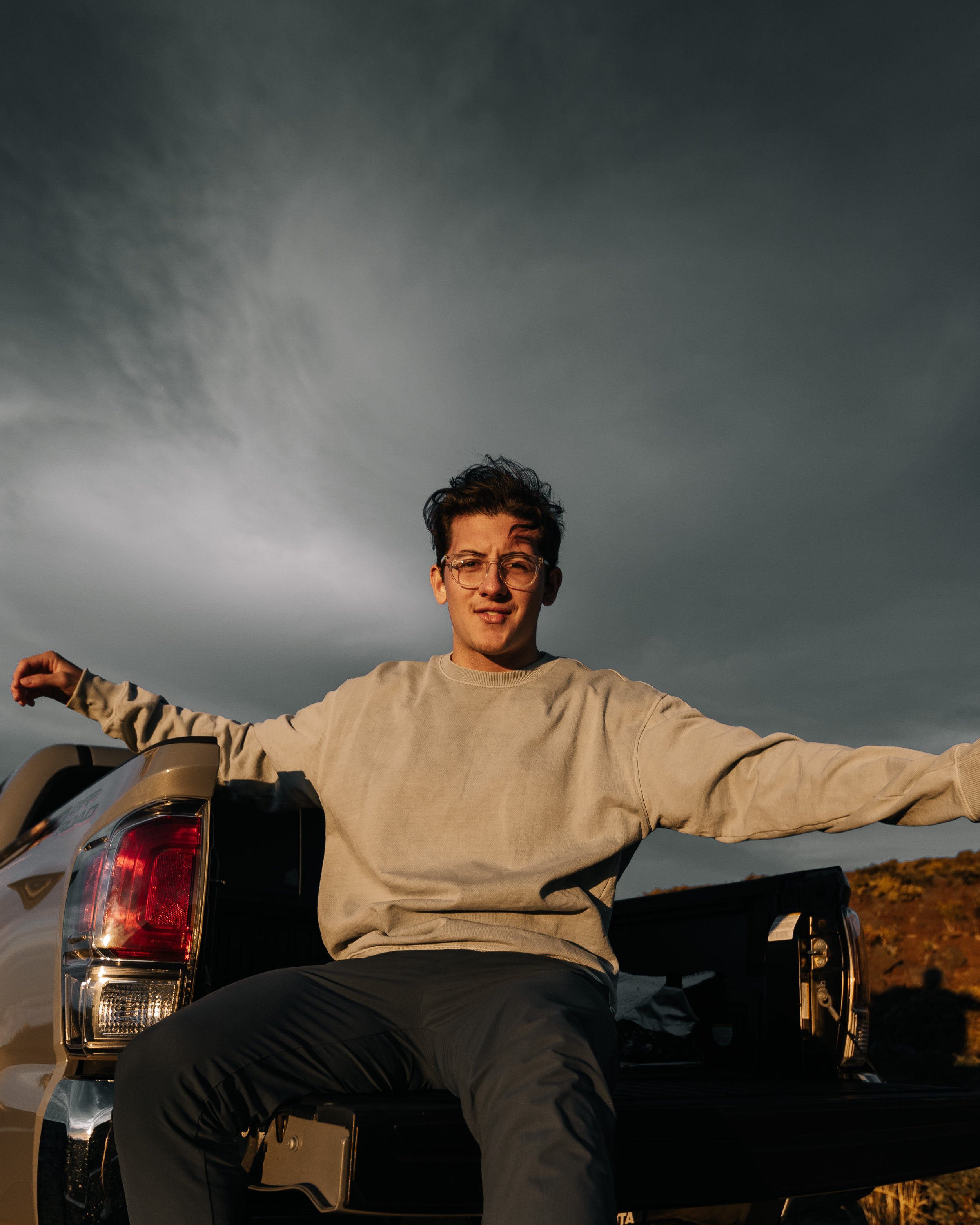Adrian Torres, owner of ADRMstudio, wearing a light beige sweatshirt and dark pants, sits on the open tailgate of a black pickup truck, with arms extended outward, against a dark, cloudy sky during sunset or sunrise.