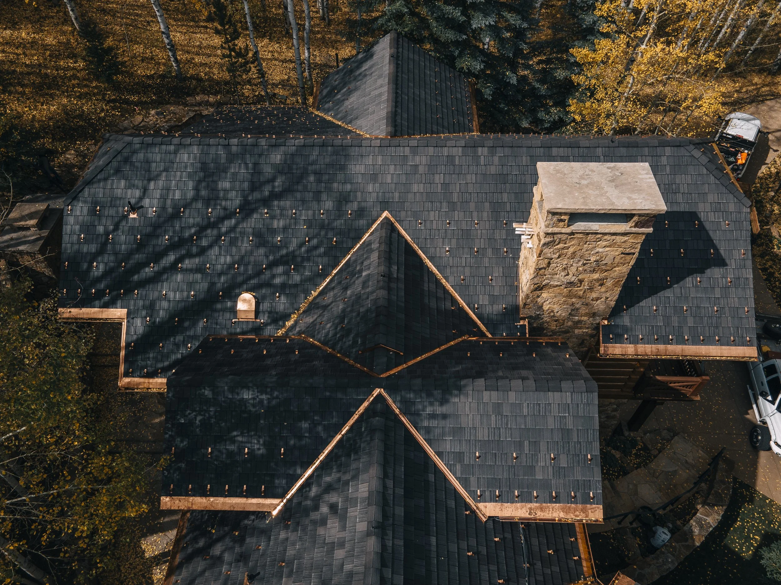 An aerial view of a house with a dark shingled roof, surrounded by trees with yellow and green leaves, and a parked vehicle visible at the bottom right corner.
