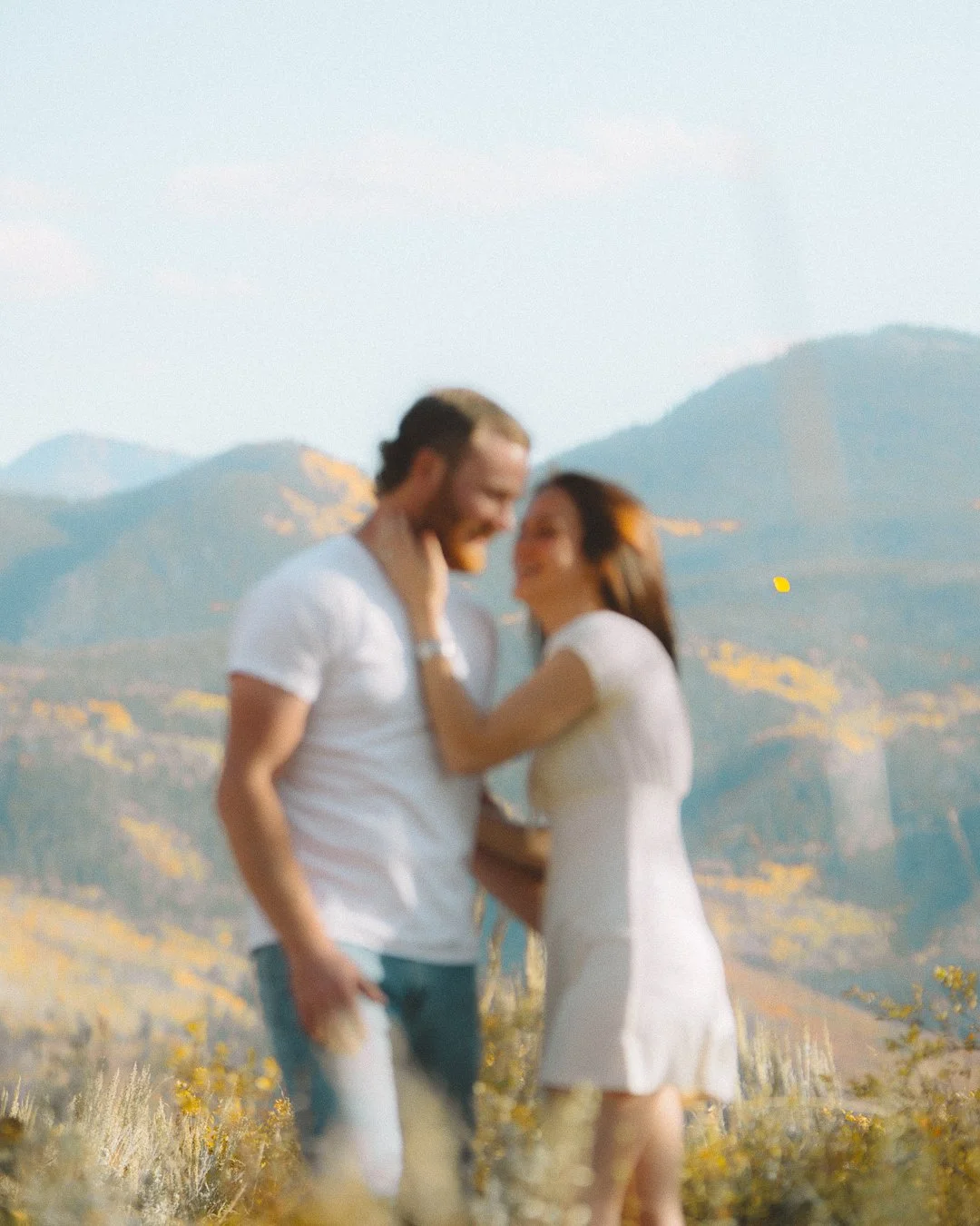 A couple standing close together in a field with mountains in the background, embracing and smiling at each other.
