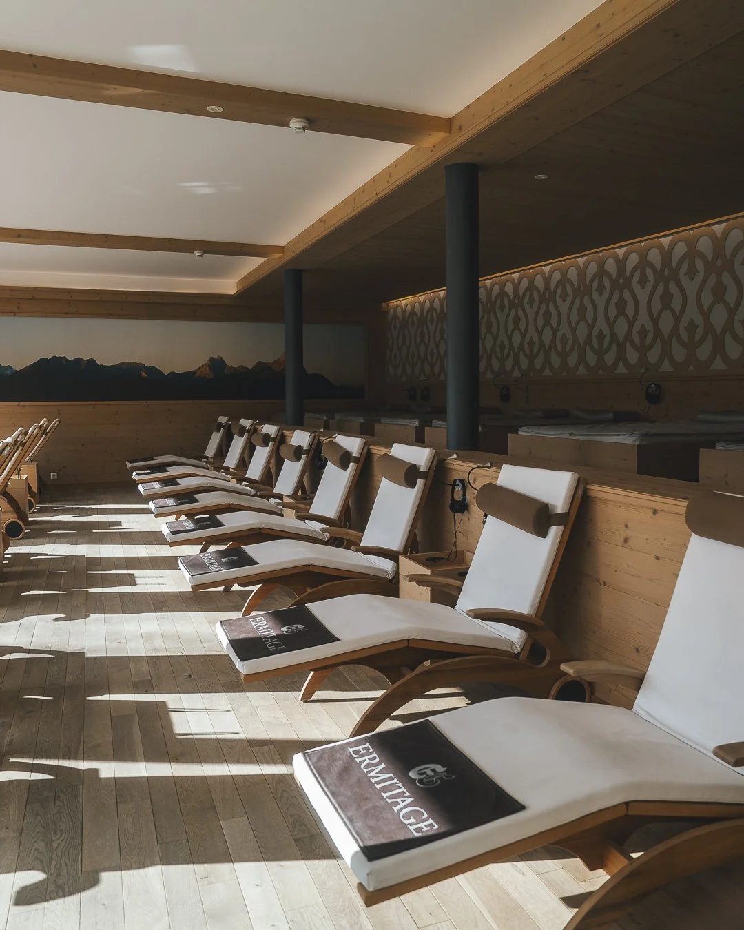 Sunlit spa relaxation lounge with wooden lounge chairs featuring white cushions and brown headrests, some with magazines, against a wooden wall with decorative patterns and a mountain mural.