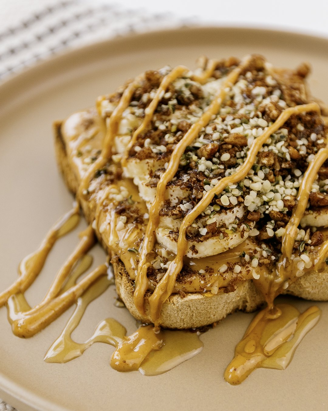 A close-up of a slice of bread topped with bananas, peanut butter, chocolate, crushed nuts, and shredded coconut, drizzled with caramel sauce on a beige plate.