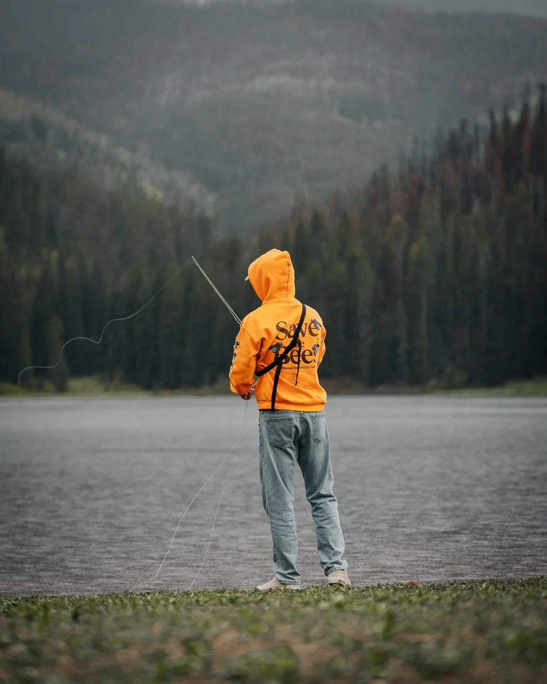 Person in orange hoodie fishing by a lake with forested mountains in the background.