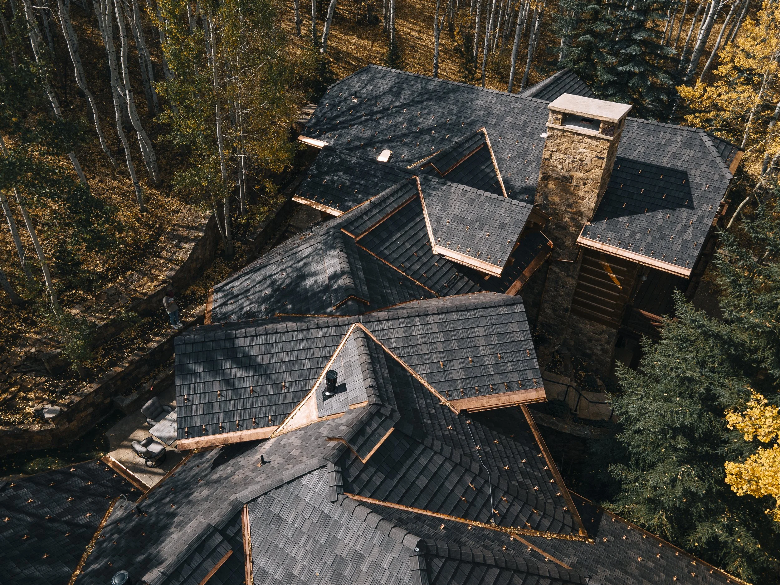Aerial view of a house with a dark gray, shingled roof surrounded by trees with autumn foliage.