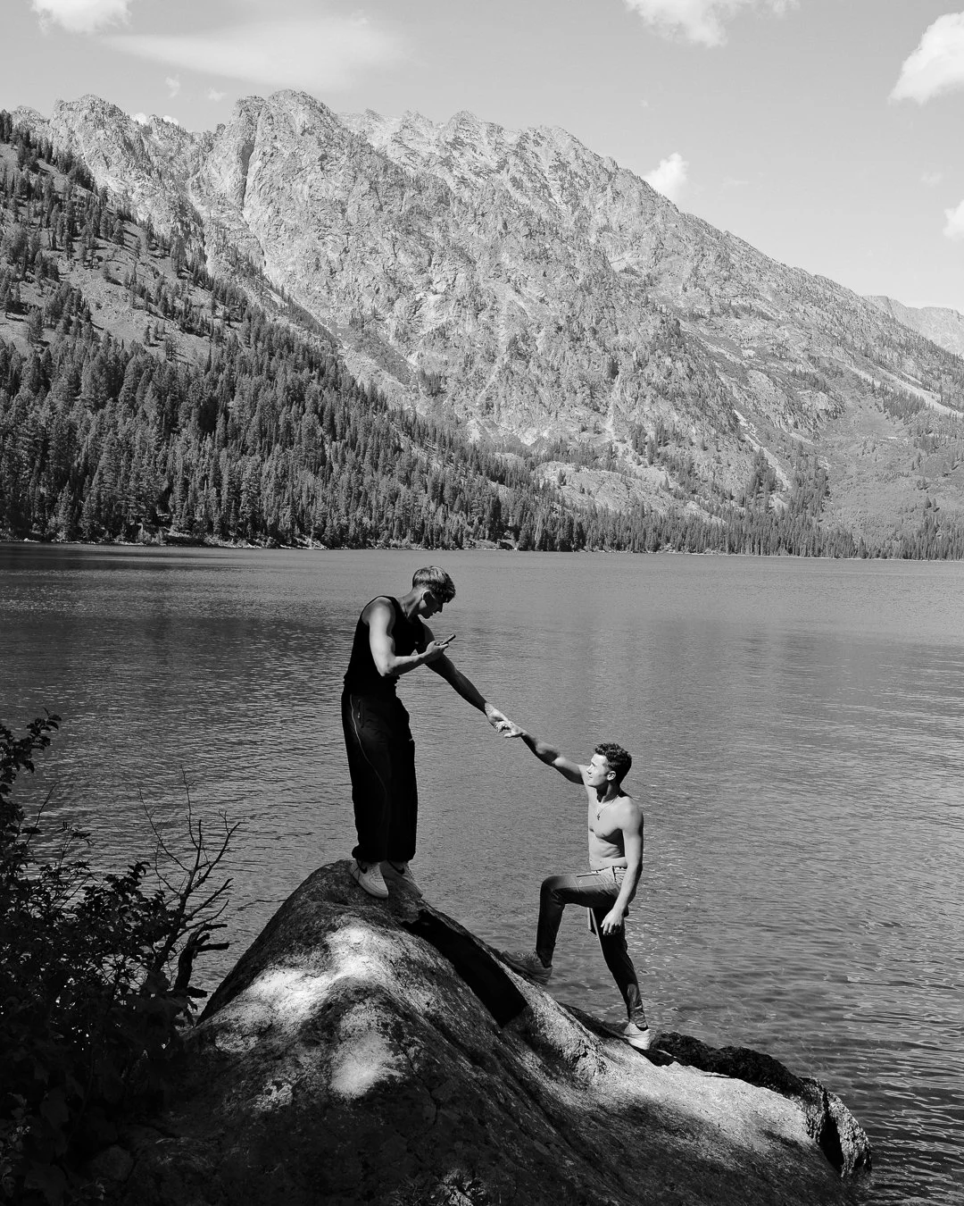 Two men on a large rock by a lake, with mountains and a forest in the background. One man is standing on the rock, reaching down and holding the hand of the other man, who is standing on the ground. Both are young men, one shirtless.