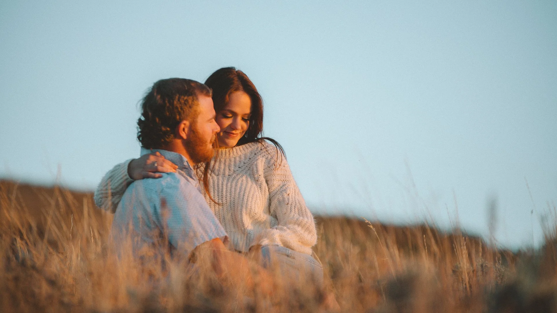 A man and woman sitting close together outdoors in a field with tall grass, sharing an intimate moment at sunset.