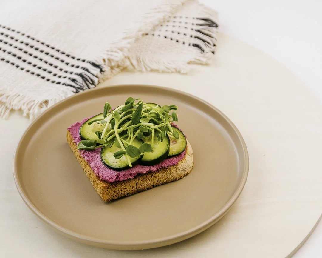 Open-faced sandwich with cucumber slices and microgreens on a beige plate, with a black and white fringed cloth in the background for Innerlight Juice.