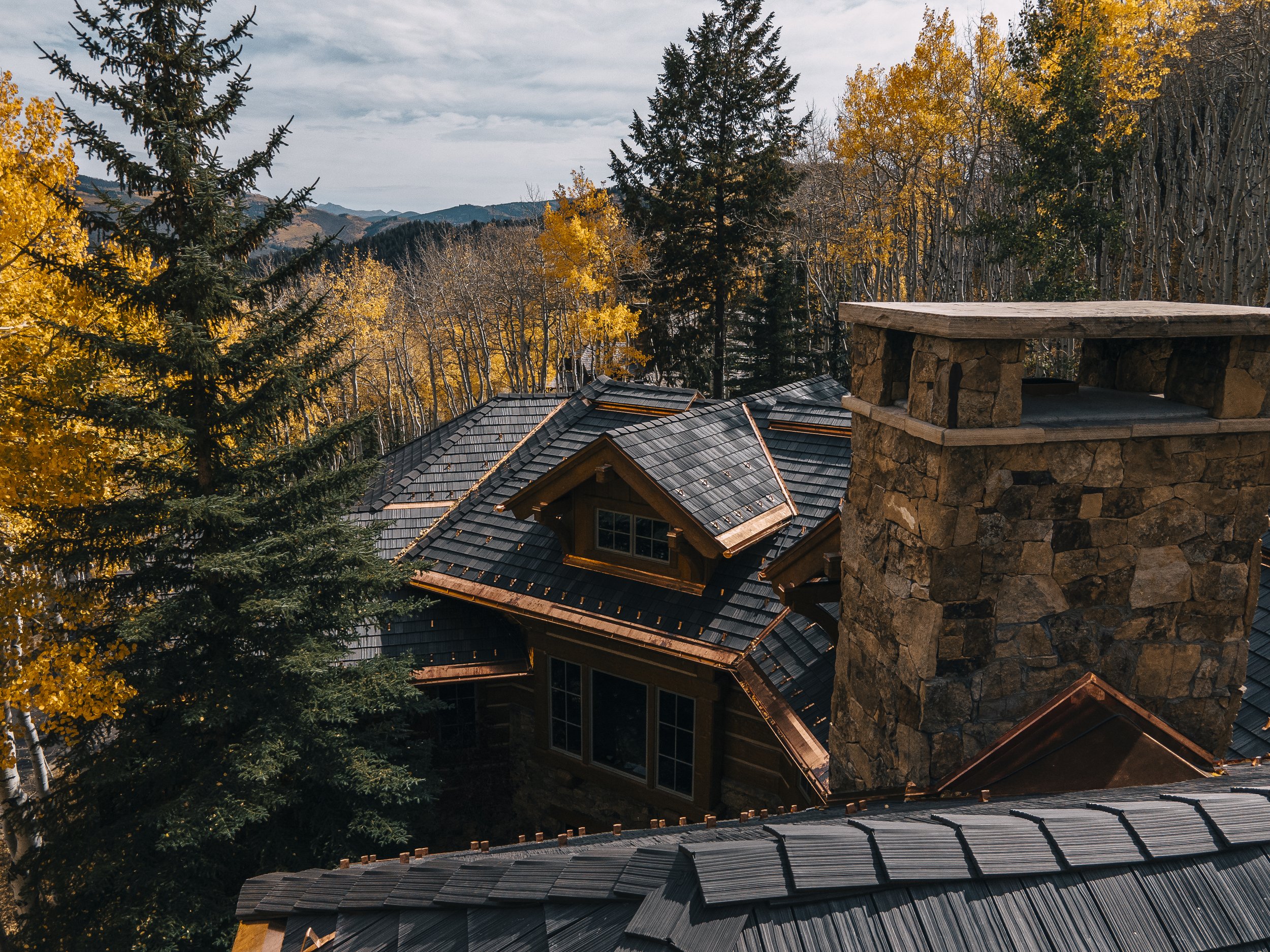 View of a mountain landscape with trees in autumn colors and rooftops of houses with dark shingles, and a stone chimney in the foreground.