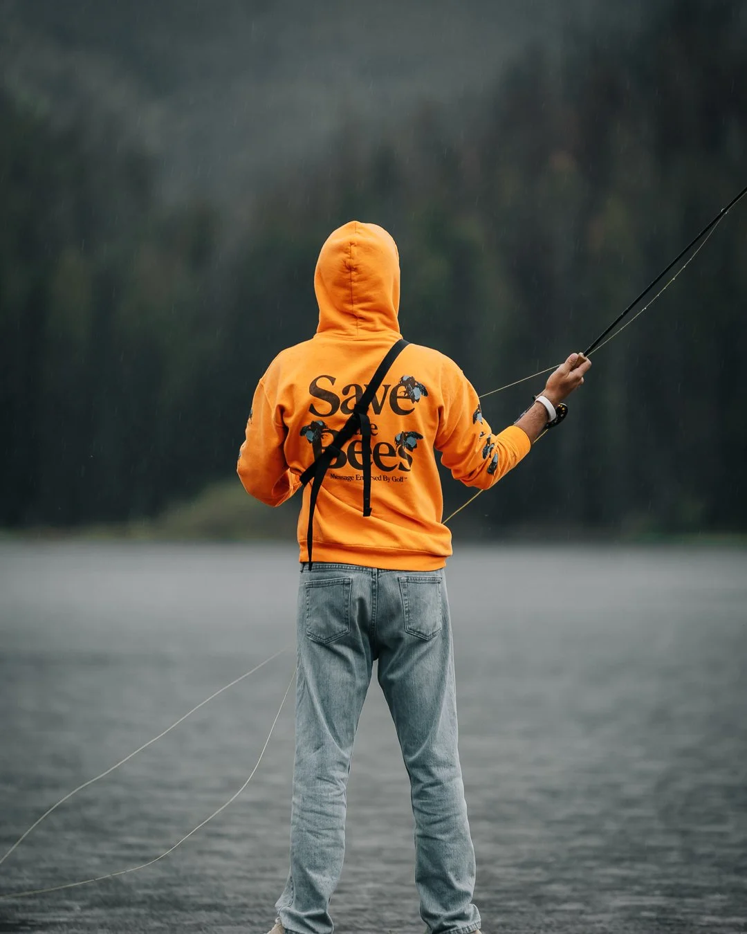 A person wearing a bright orange hoodie with the words 'Save the Roses' on the back, standing by a body of water, fishing during a rainy day.