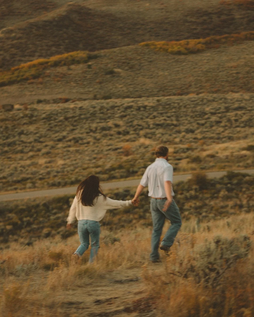 A couple walking hand in hand through a grassy field in a hilly landscape during sunset.