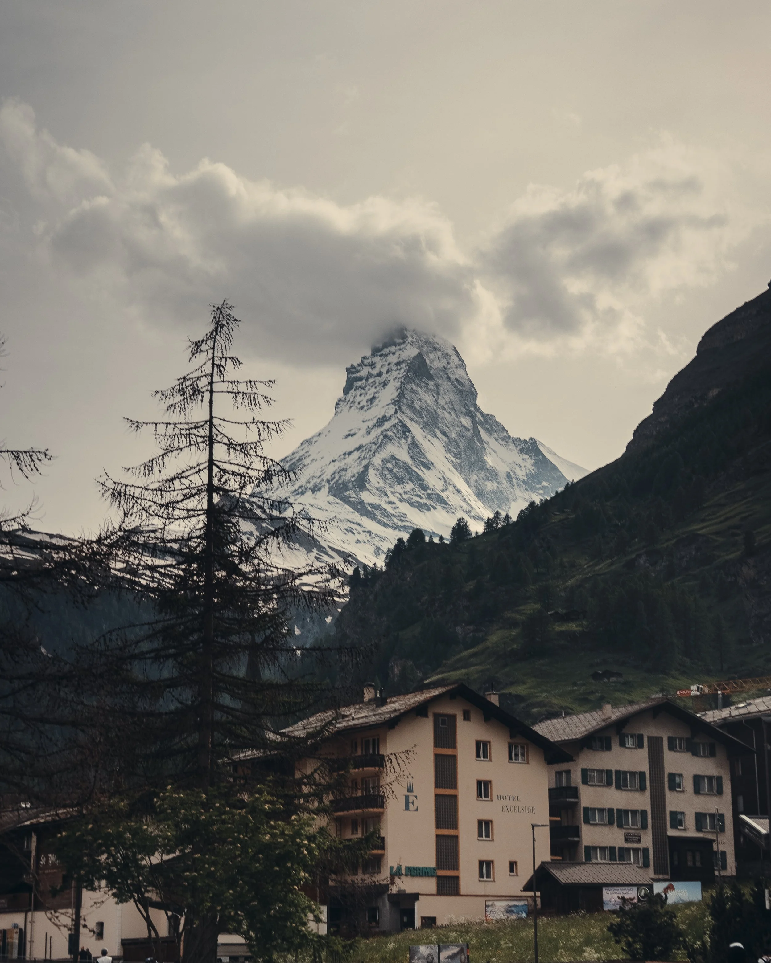 The Matterhorn, a snow-capped mountain peak, partially obscured by clouds, stands behind a small town with hotels and trees in the foreground.