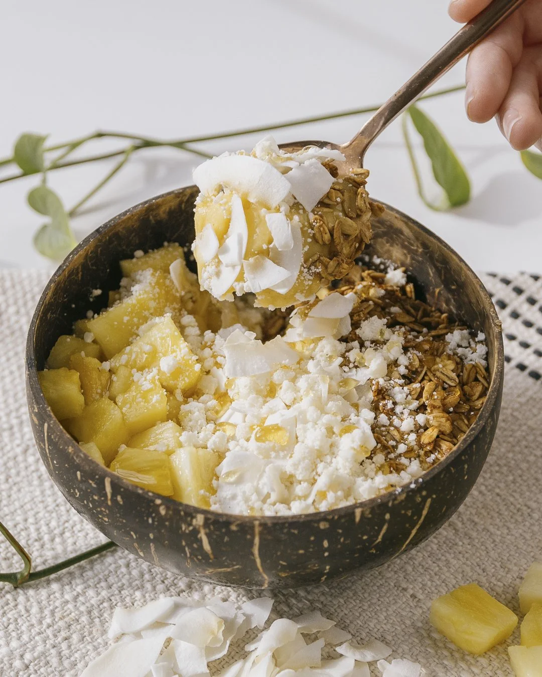 A black coconut bowl filled with pineapple chunks, granola, shredded coconut, and white crumbles, with a spoon lifting a scoop from the bowl.