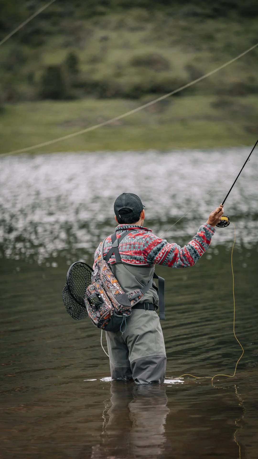 A person standing in a shallow river, fishing with a fishing rod. They are wearing a cap, a patterned jacket, and carrying a backpack and a fishing net.