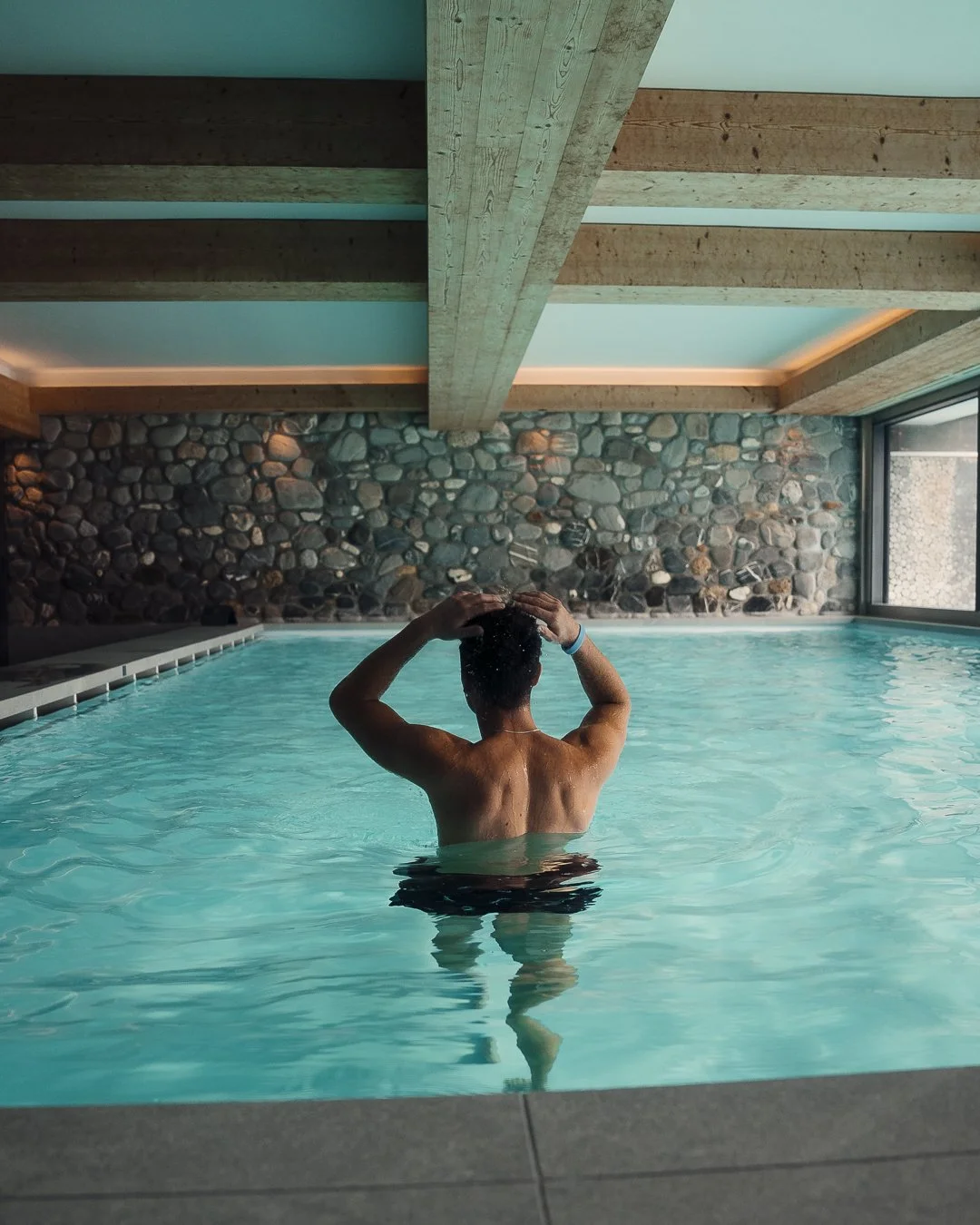 Ermitage Wellness & Spa Hotel in Gstaad. A person is standing in an indoor swimming pool with their back to the camera, adjusting their hair. The pool area features a stone wall, wooden ceiling beams, and a large window letting in natural light.