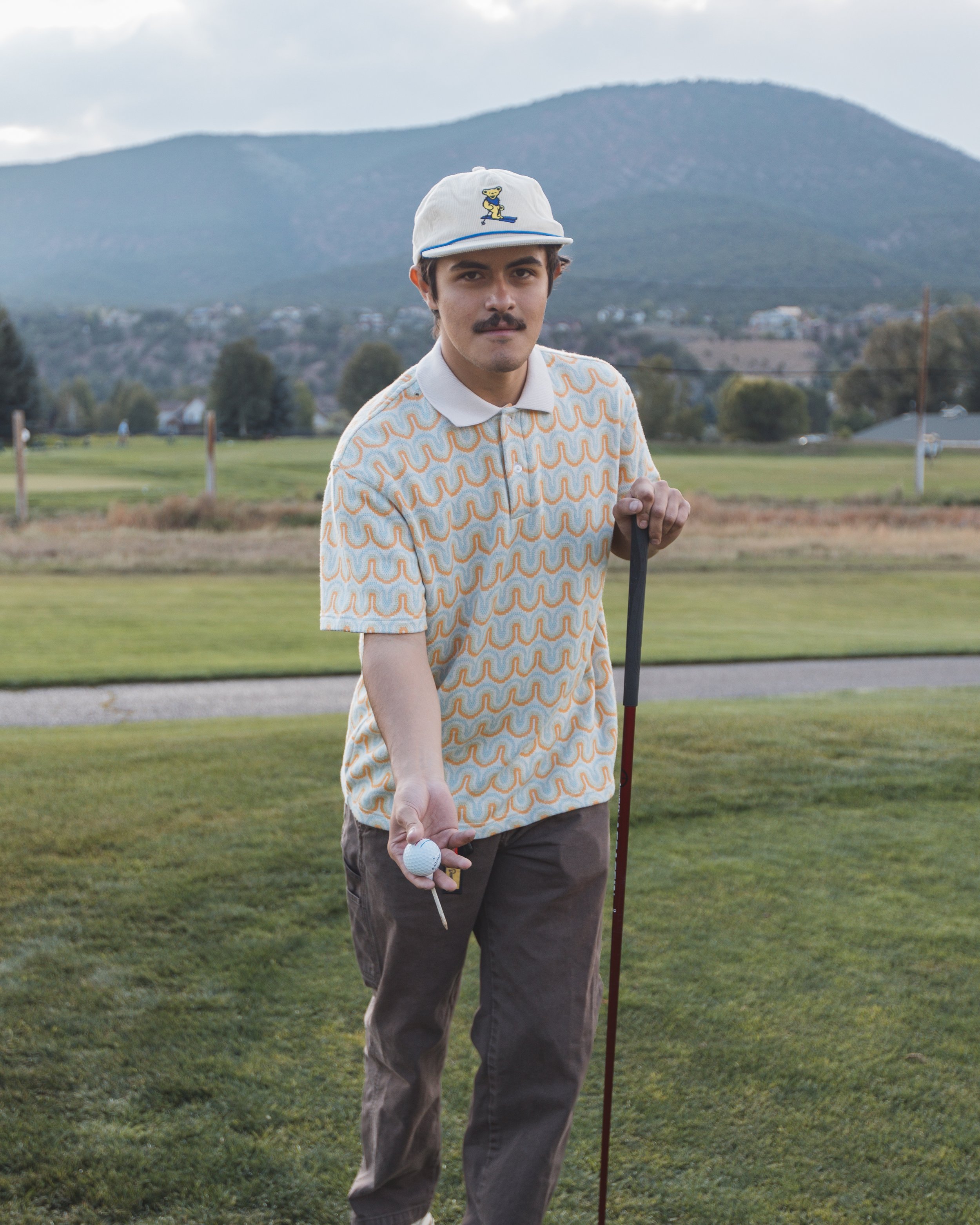A man wearing a colorful patterned polo shirt and brown pants is standing on a golf course, holding a golf club and a golf ball, with in the background trees, houses, and mountains under a cloudy sky.