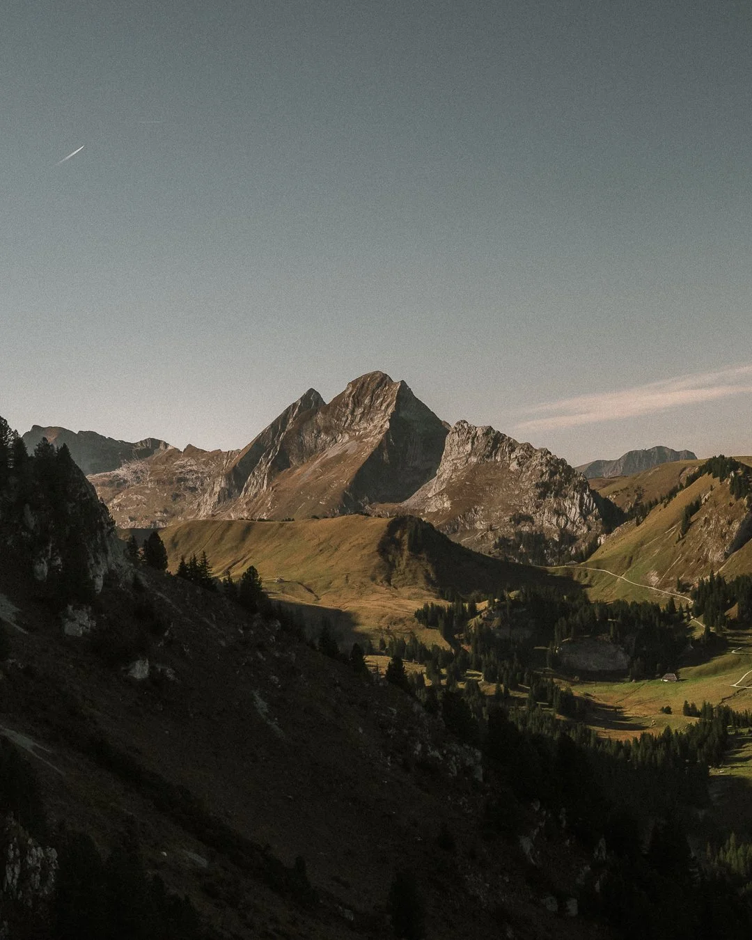 Scenic mountain landscape with rugged peaks, green valleys, and scattered trees under a clear sky.