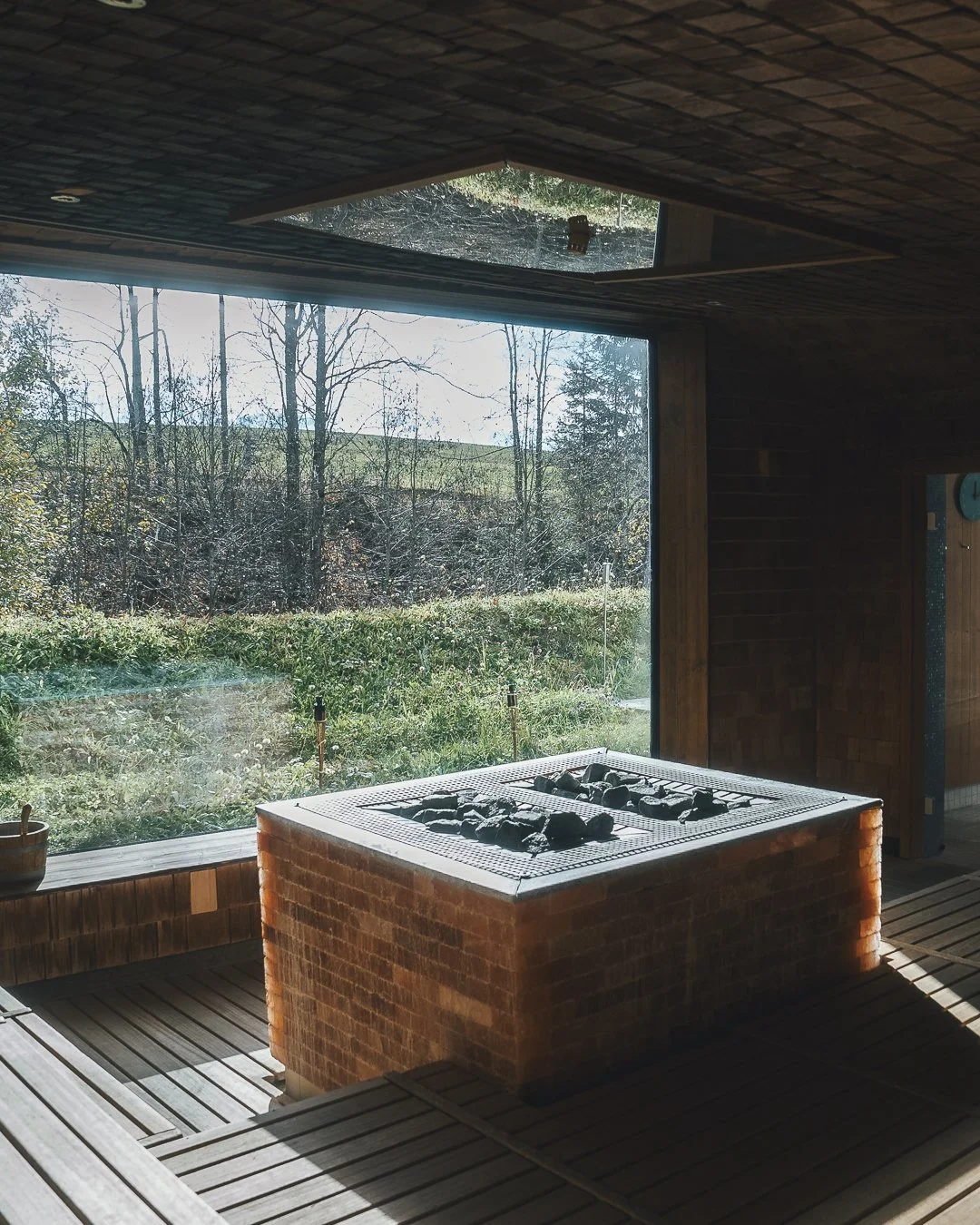 Indoor space with a brick firepit on a wooden deck, large window showing trees and outdoors, and a ceiling cutout with a view of the sky.