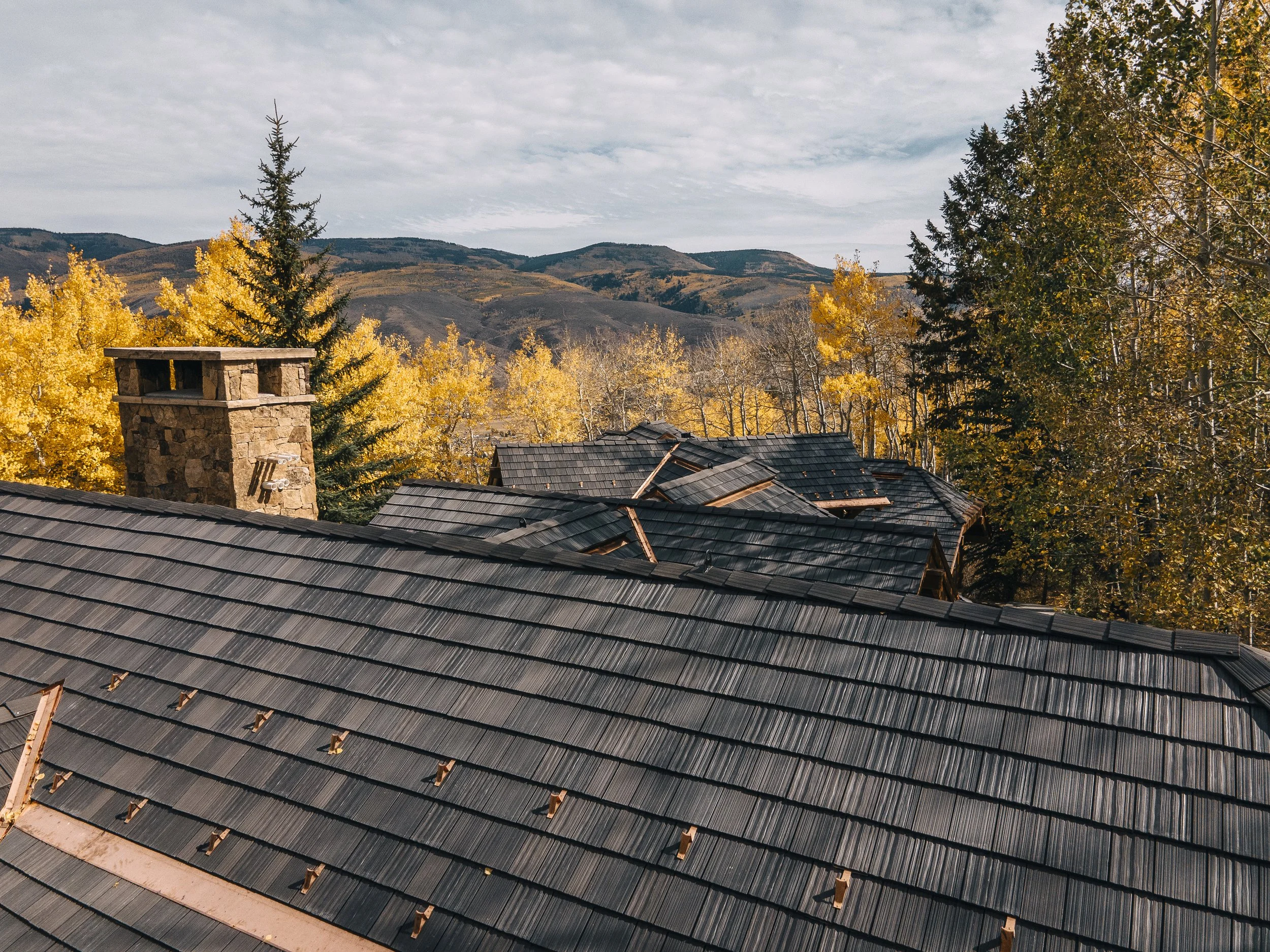 A rooftop with black shingles surrounded by trees with yellow and green fall foliage and a stone chimney, set against a backdrop of mountains and a cloudy sky.