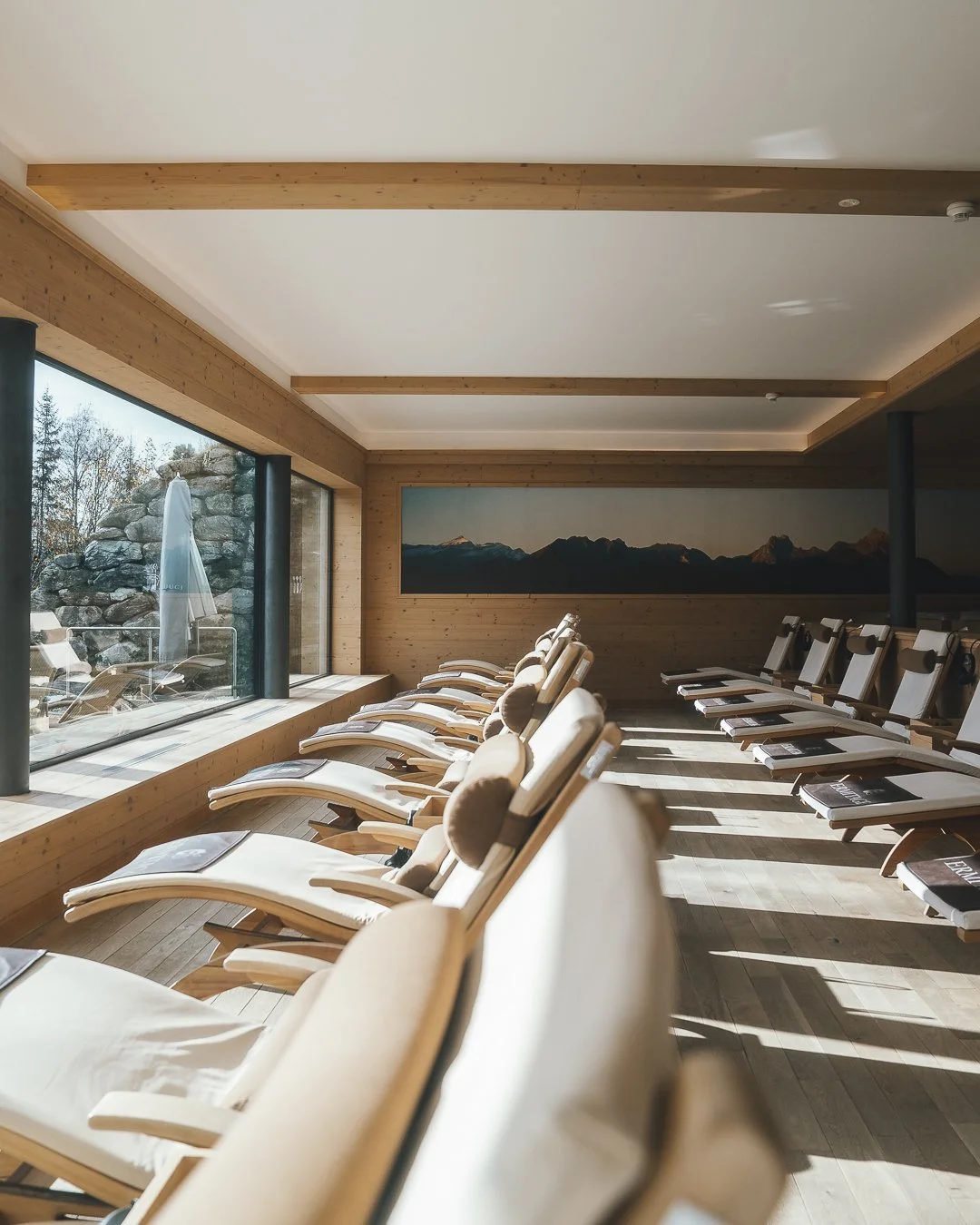 Spa or relaxation room with beige lounge chairs near large glass windows showing outdoor scenery and a mountain painting on the wall.
