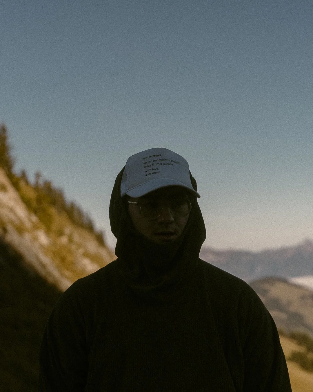 A person wearing a black hoodie and a cap with text, standing outdoors during dusk with mountains in the background.