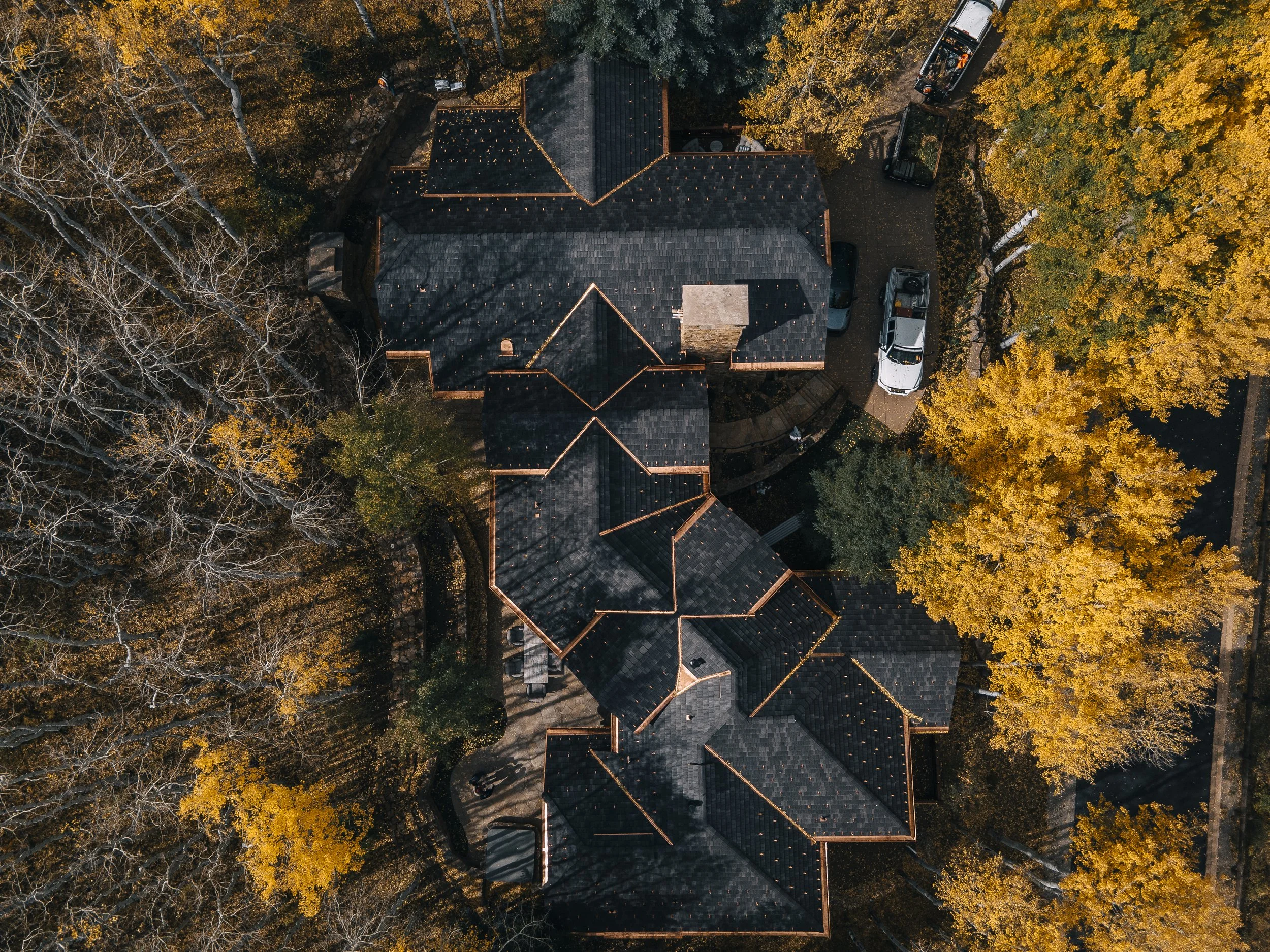 Aerial view of a house surrounded by trees with yellow and orange fall foliage, cars parked in the driveway, and a dark shingled roof.