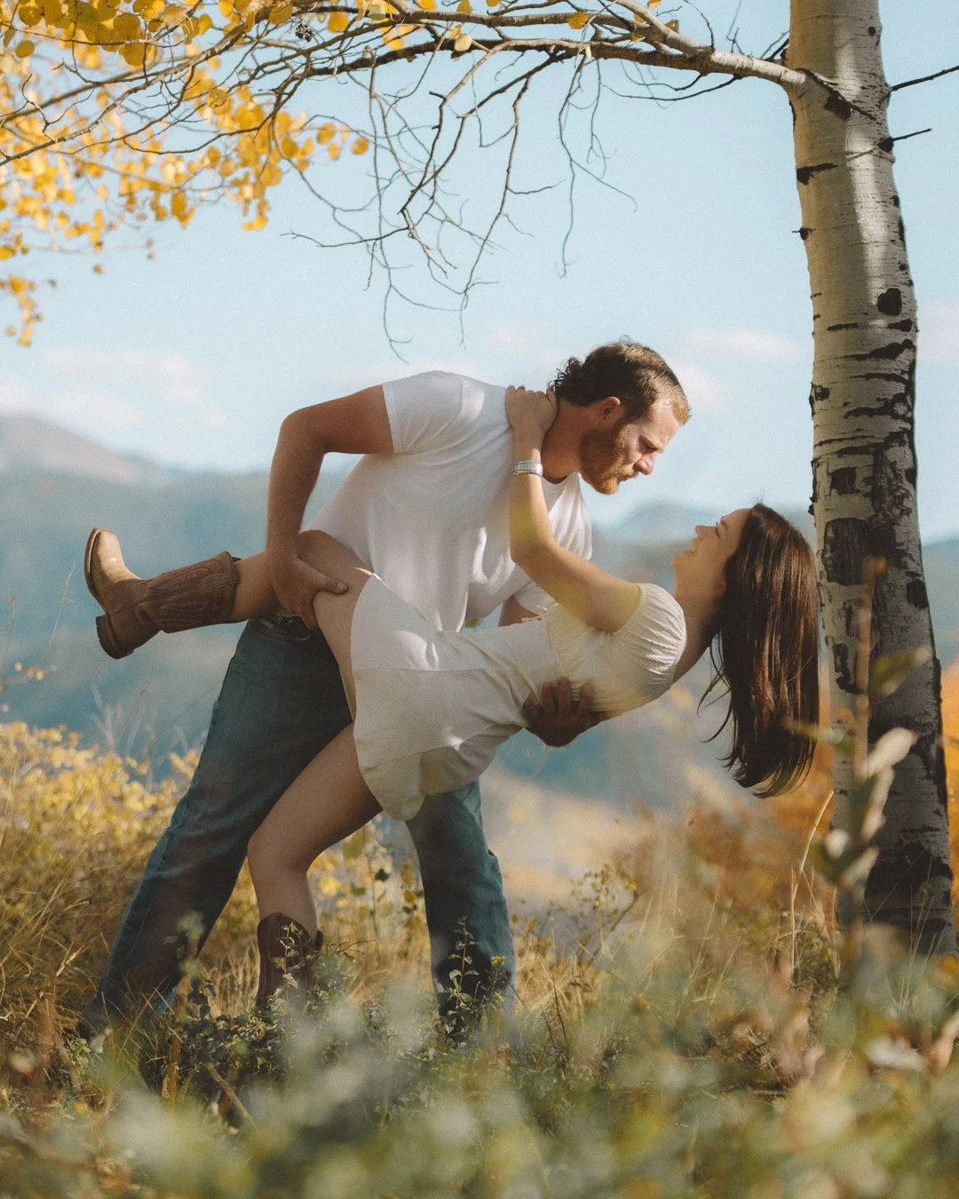 A man holding a woman in a dip pose outdoors during fall, near a tree with yellow leaves, with mountains in the background.