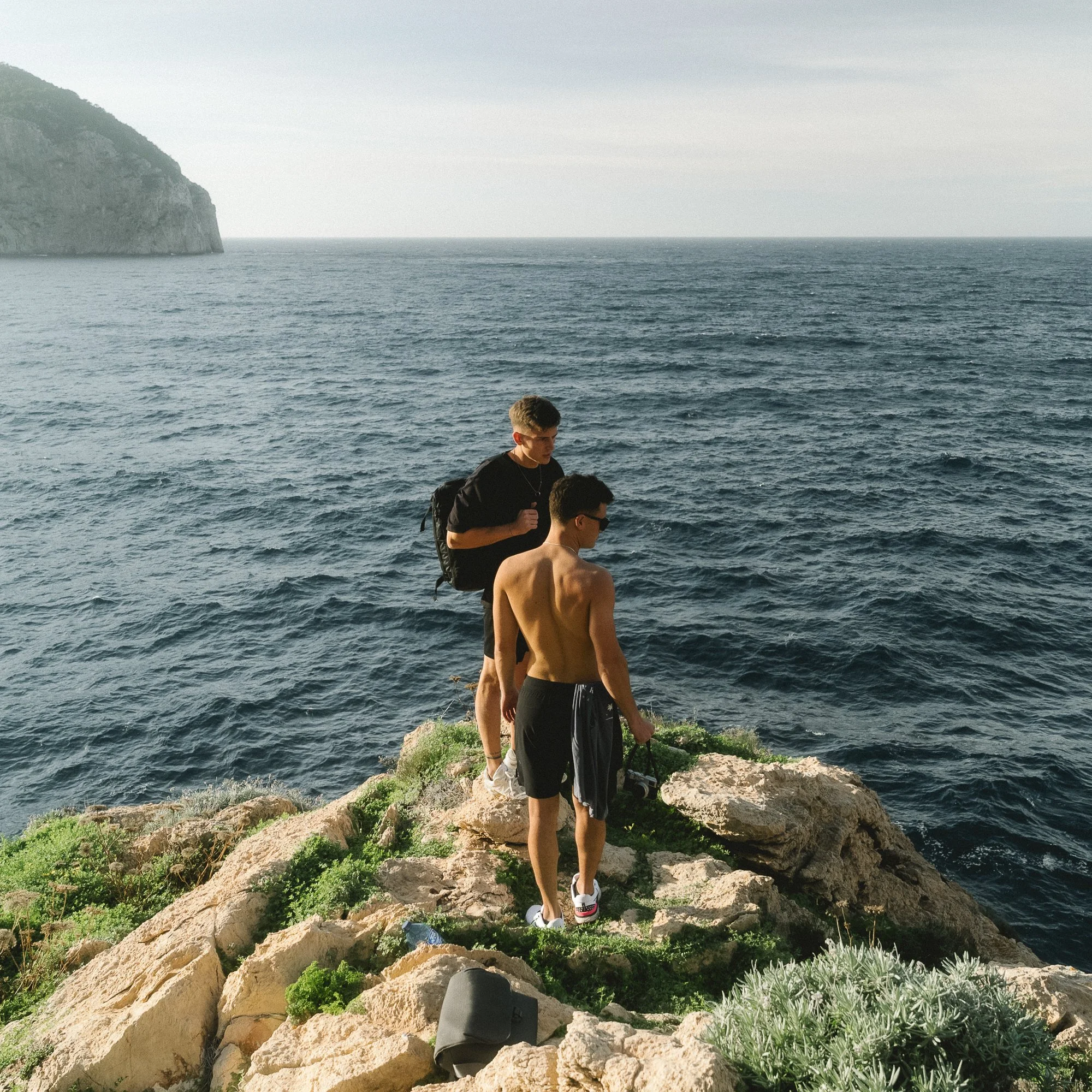 Three young men hiking along a rocky coastal trail with the ocean in the background. Two of them are shirtless and the other has a backpack, and a camera is visible on the ground. A hillside with greenery is on the left, and the sea stretches to the 