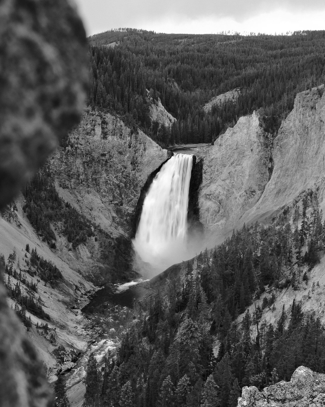 Black and white photo of a waterfall pouring into a canyon surrounded by dense forest and mountains.